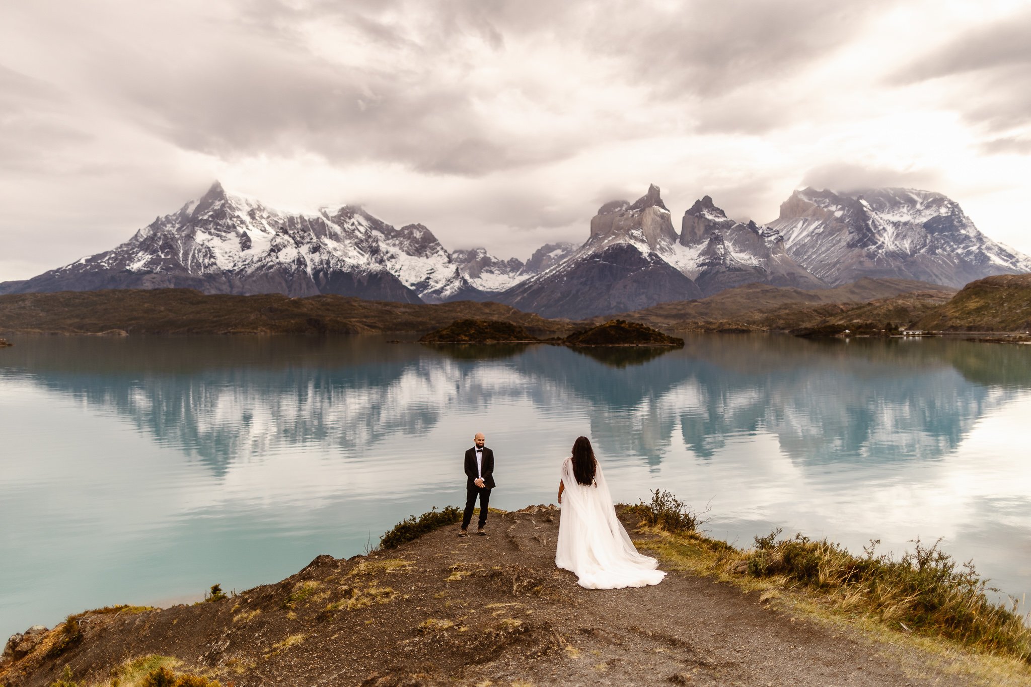 Right on the edge of the turquoise waters of Torres del Paine, the groom stands at the end while watching his bride walk towards him in her flowy wedding gown. The jagged peaks fill the background on a moody, cloudy day.