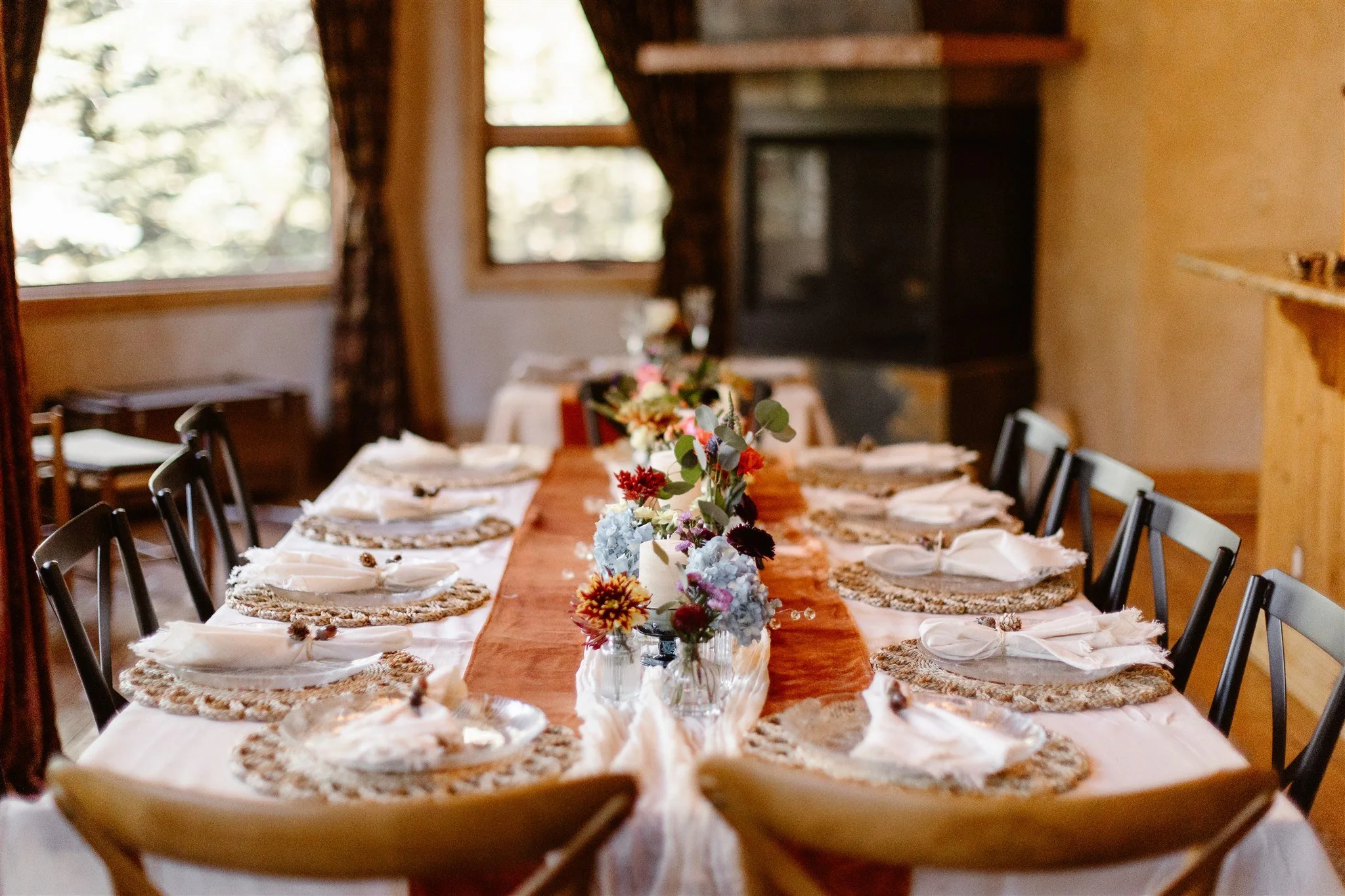 The table setting for the intimate wedding reception in an rental home. The table is decorated with an orange cheesecloth table runner, braided placemats under the clear glass plates, and mini flower vases filled with blue, purple, and orange flowers