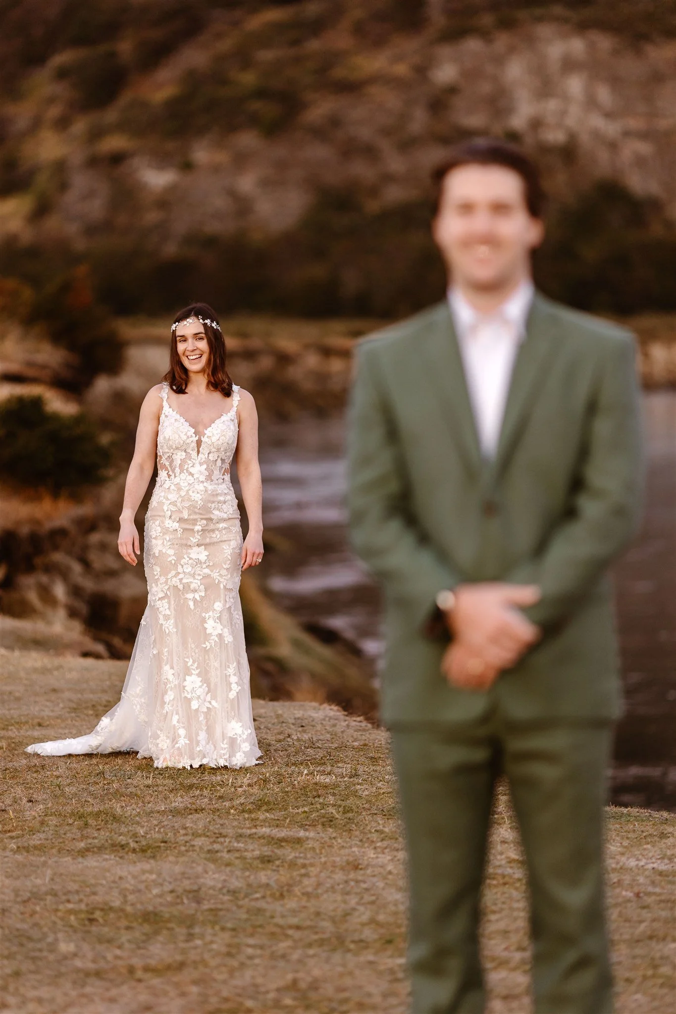 During a sunrise first look in Patagonia, the bride stands behind the groom with a bright smile on her face as she prepares to walk to her fiance, who is out of focus to the right, in front of the camera, wearing a green suit.
