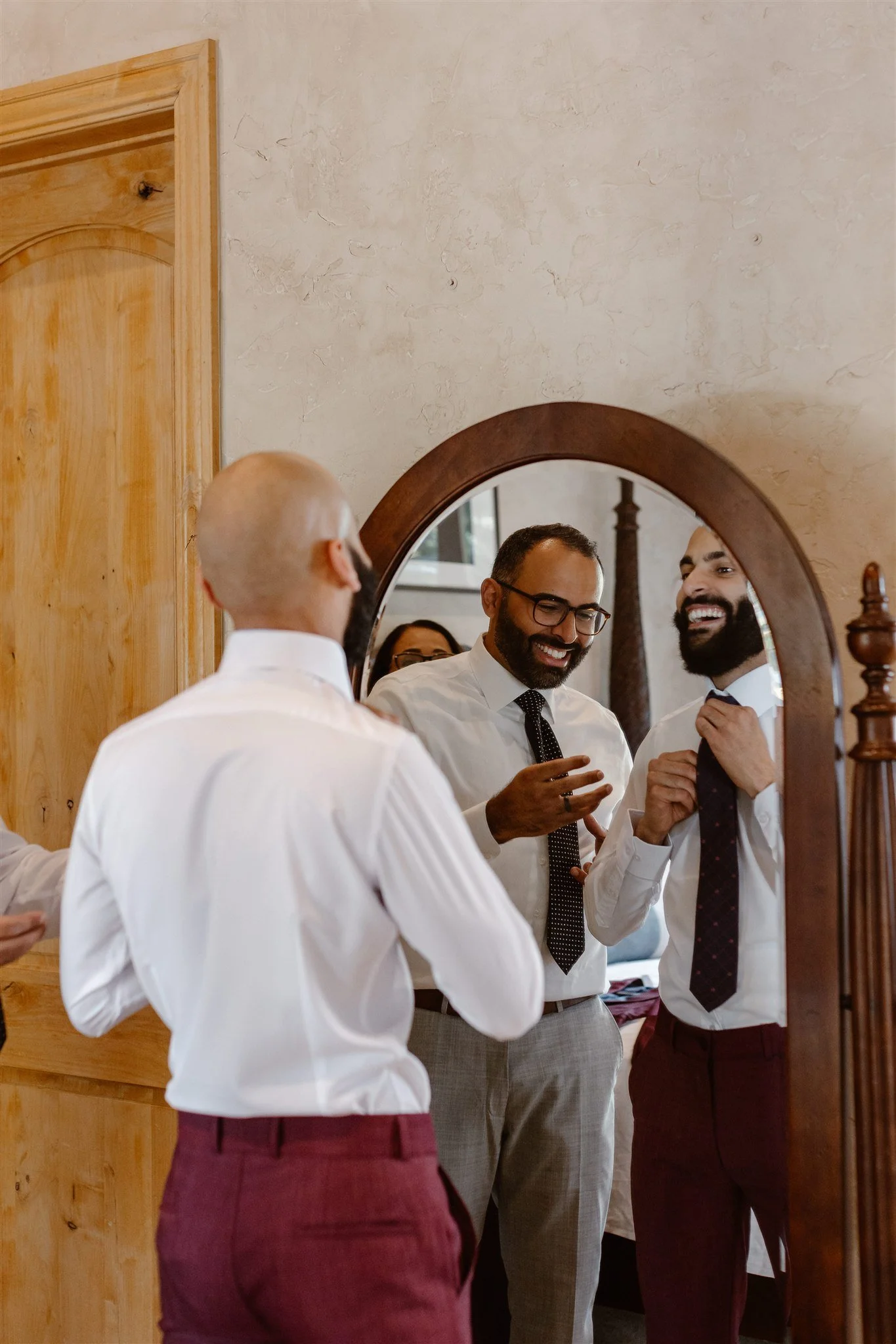 The groom putting on his tie in the mirror while laughing joyfully with his friends and family beside him. He's tightening a red tie over his crisp white shirt and maroon colored dress pants.