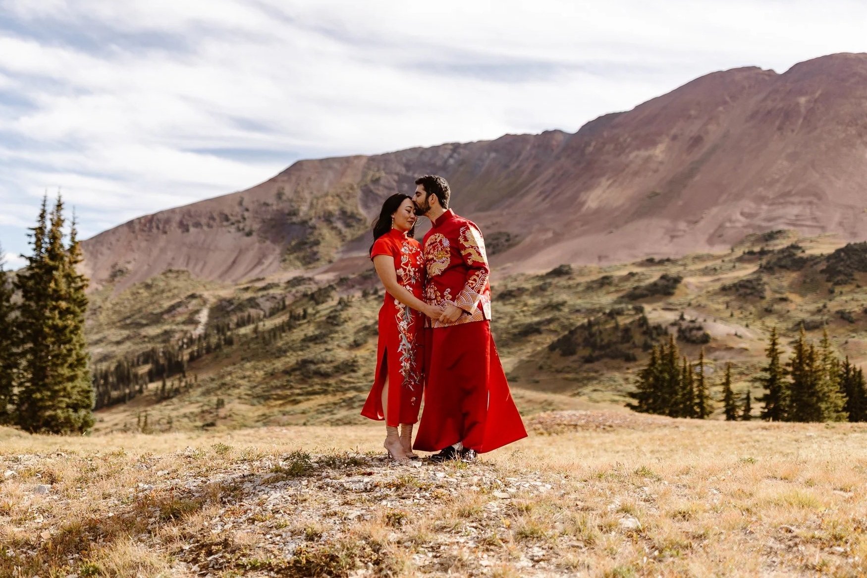 During their elopement portraits in the mountains of Crested Butte, an interracial couple share a tender moment of stillness while in their red traditional Chinese wedding outfits. The groom kisses his bride's forehead as the rest of the world pauses