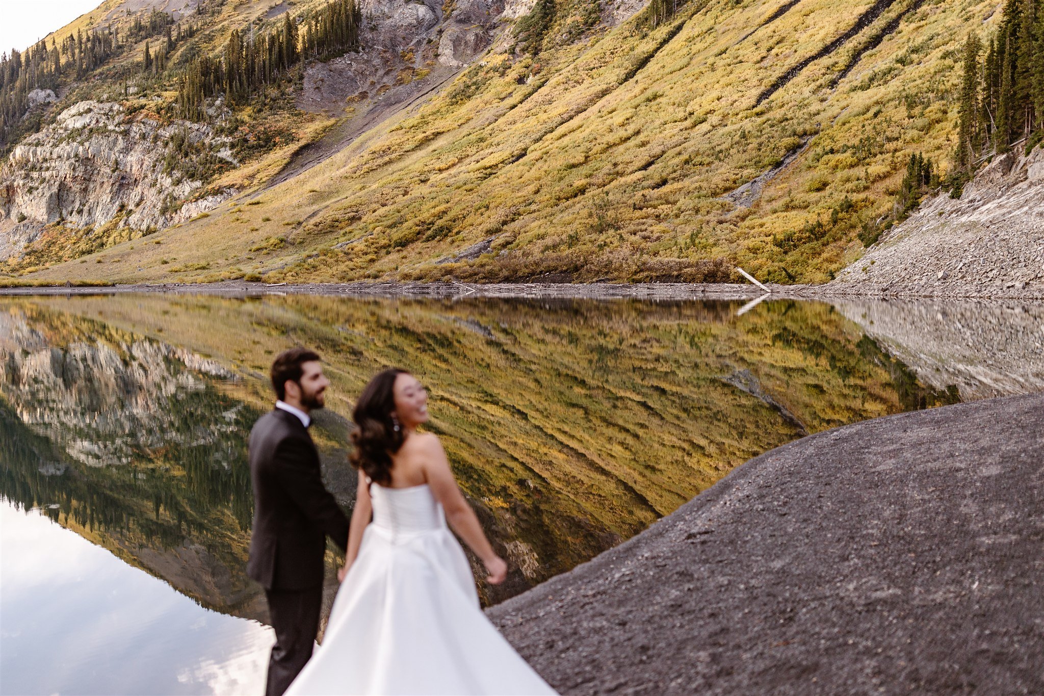 This image is focused on the mountains and lake's reflection, with the smiling couples blurred out in the foreground. The background pops with shades of green and yellow, the perfect contrast against the dark color of the lake's shore and rocks.
