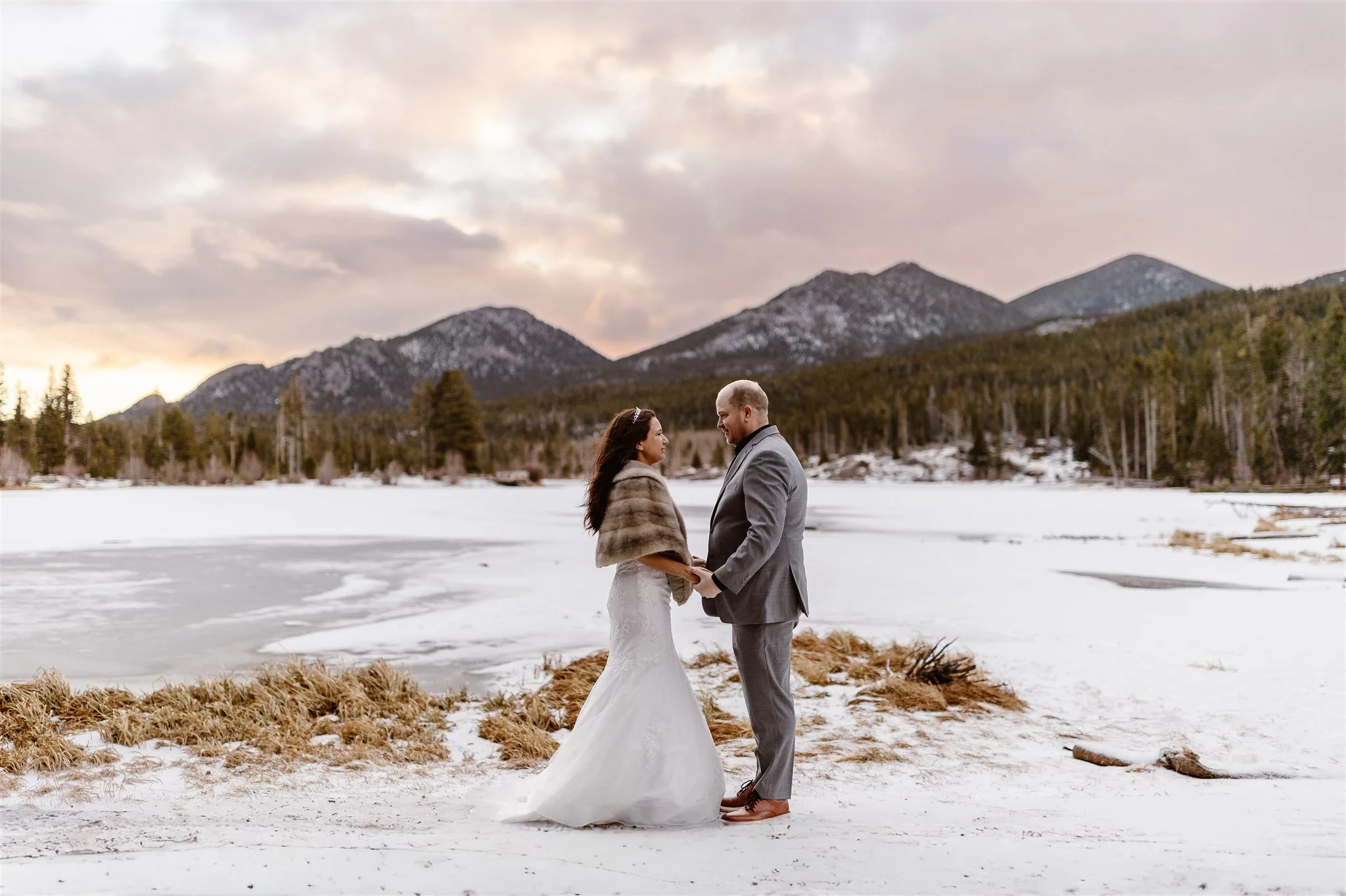 During their sunrise elopement in Rocky Mountain National Park, the bride stands to the left in her mermaid wedding dress and fur shawl, while the groom stands to the right in his grey suit. The clouds are softly lit in pink and yellow above them.