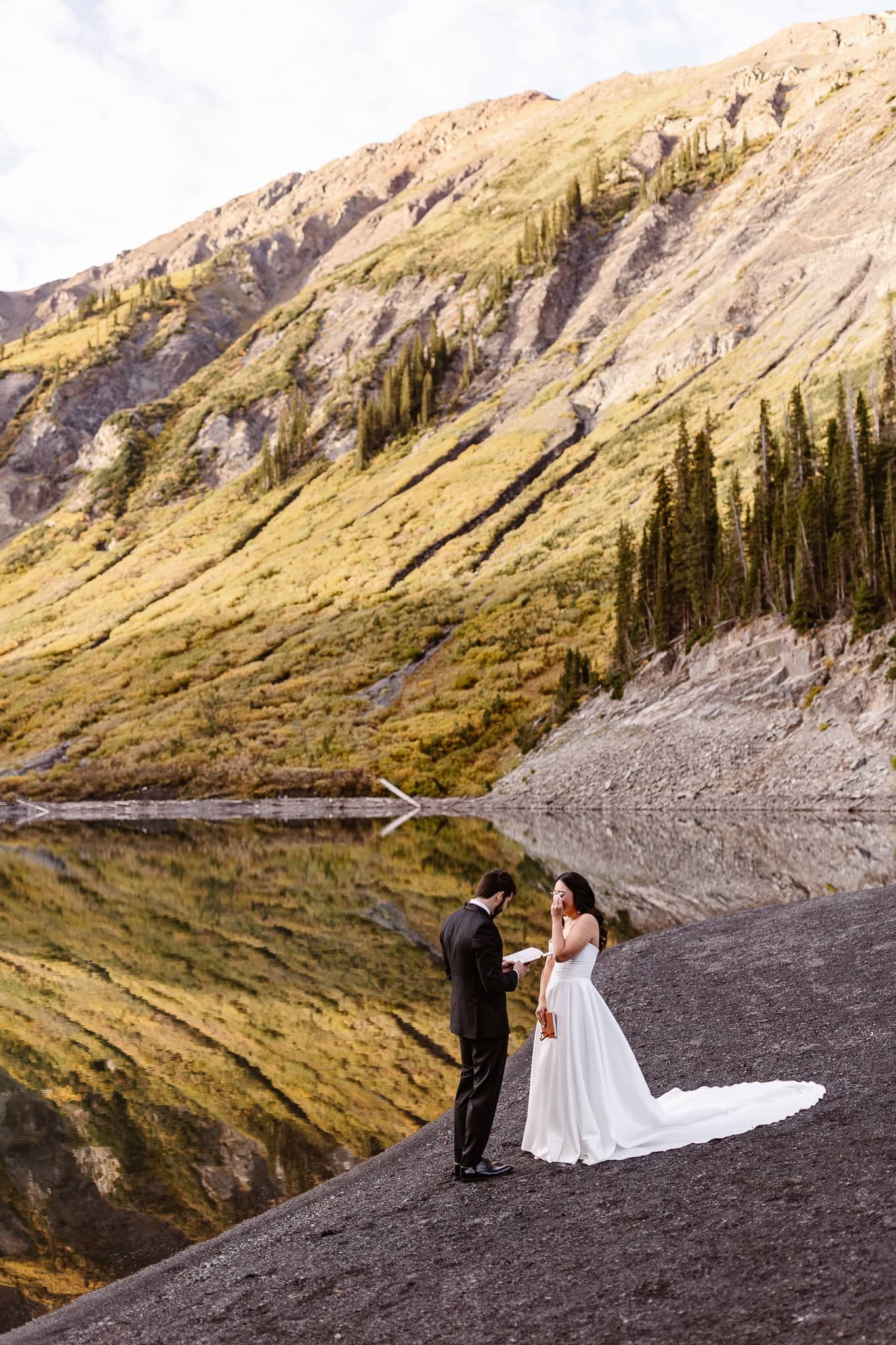 As the groom reads his vows on the edge of the lake, the bride wipes her tears while holding her vow book in the other hand.