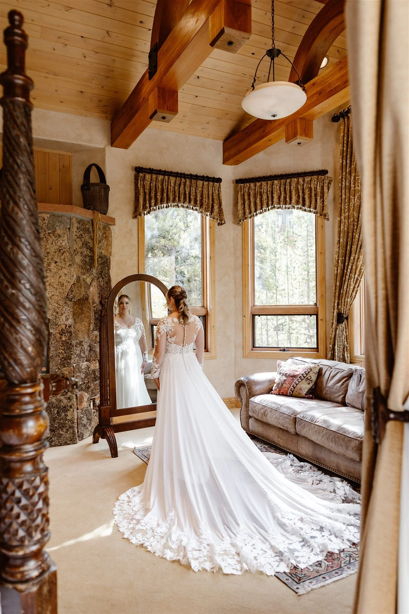 The bride looks in the mirror as she takes in her wedding-day look on a calm afternoon in Breckenridge. This photo shows the full extent of the gorgeous lace train of the light and airy gown and the somewhat exposed back of her dress.