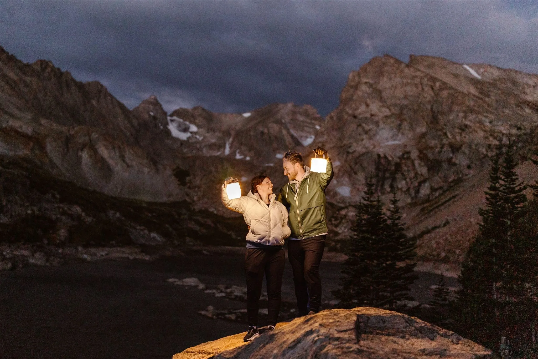 During their sunrise elopement, the bride and groom stand on a rock before the towering mountains at Lake Isabelle, still dressed in their hiking gear and holding lanterns while smiling at each other, waiting for the sunrise.