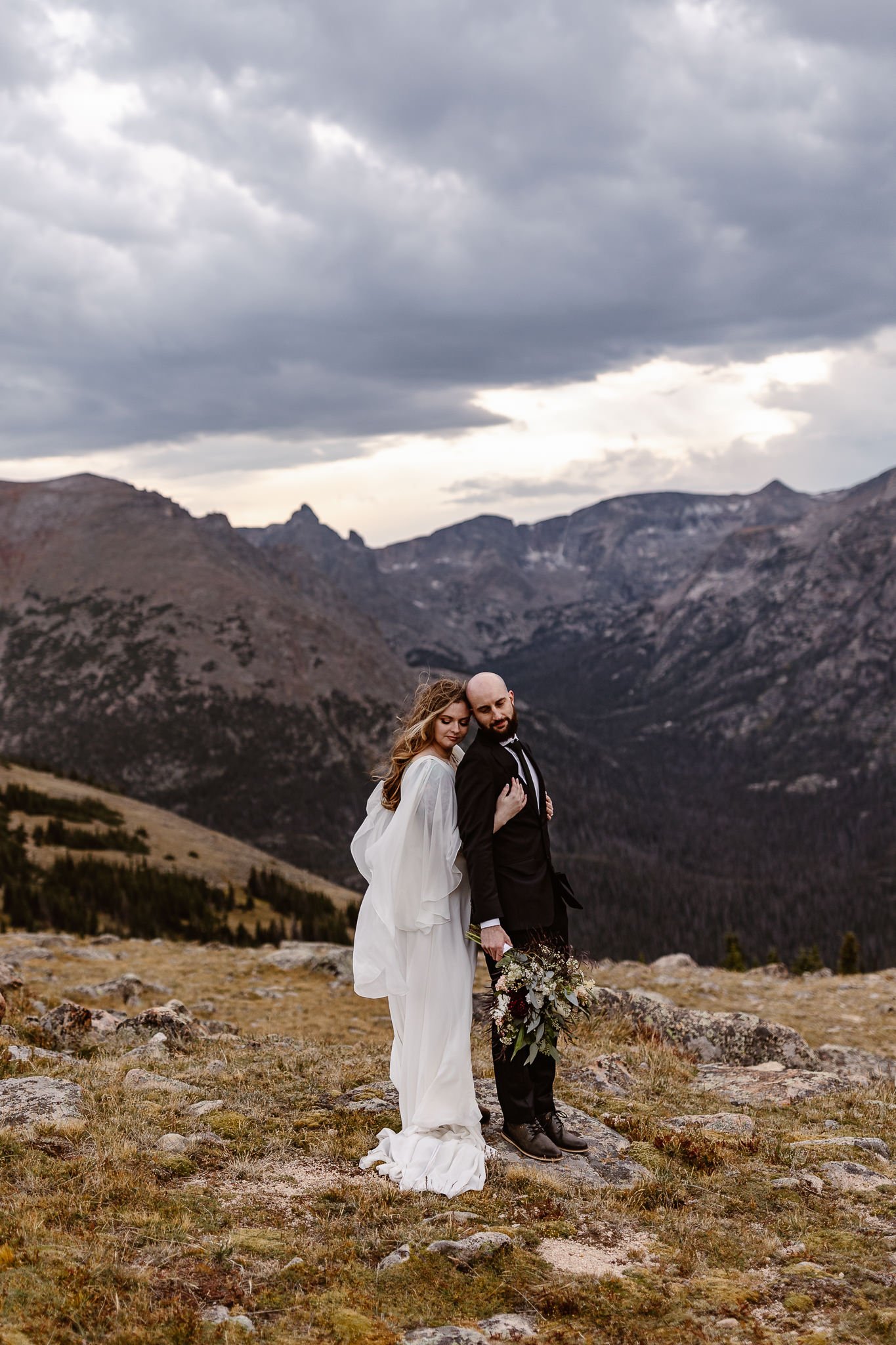 A vertical image of the newlyweds closing their eyes and soaking in the moment amongst the mountains on this epic day. The bride is holding her husband from behind, both hands on this chest/stomach, as he leans back towards her with her bouquet.