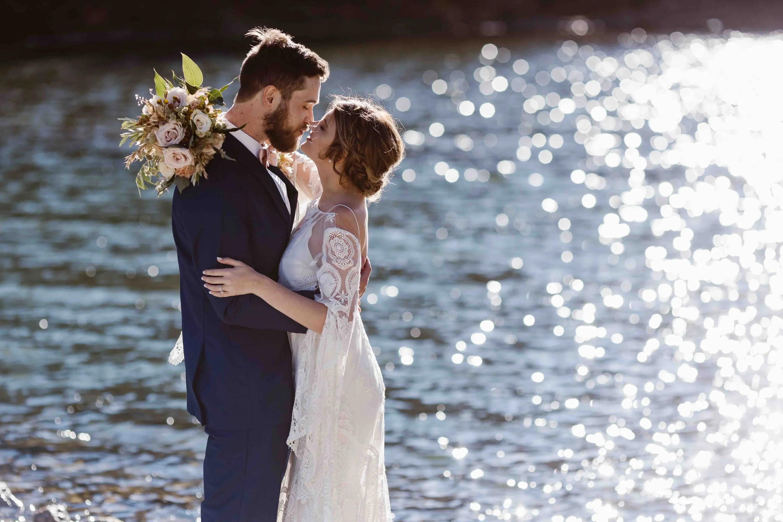 Bride and groom embrace and almost kiss standing at the edge of a shimmering lake