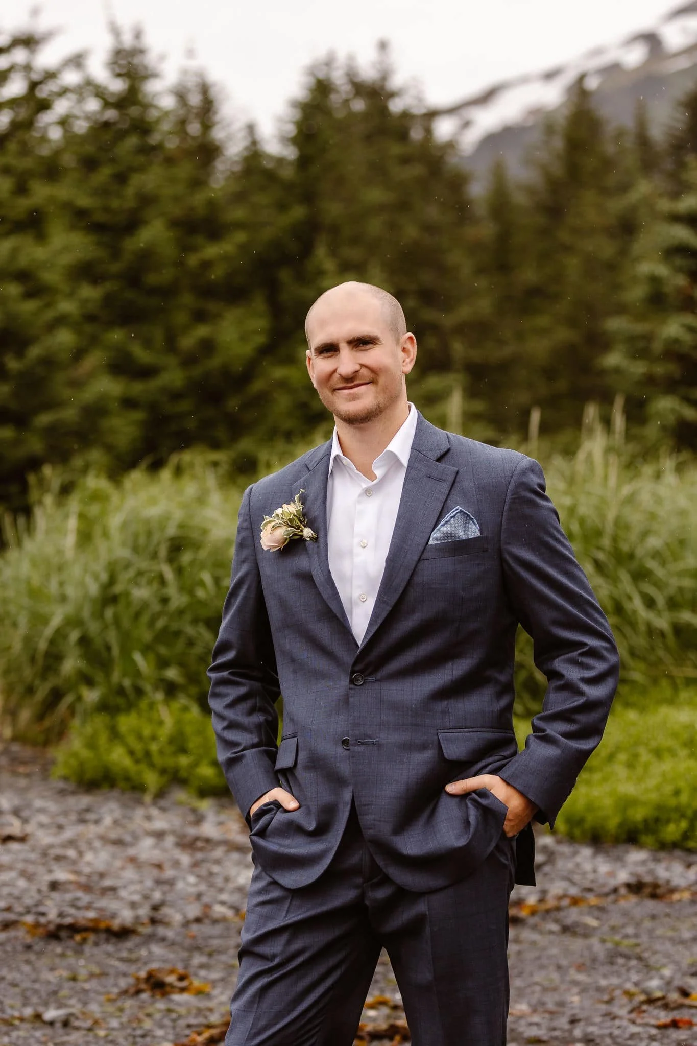 On his way to the meeting spot for their first look, the groom stands at the edge of the beach where the grass and trees meet the sand, standing with a friendly smile with his hands in his pockets.