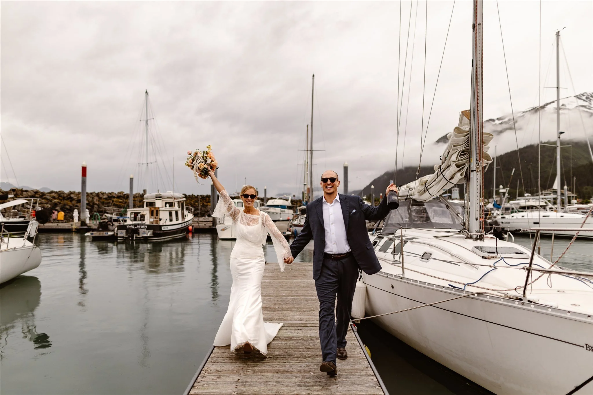A happily newly-married couple hold hands as they walk down the pier, the bride holding her bouquet in the air and the groom holding up a bottle of champagne.