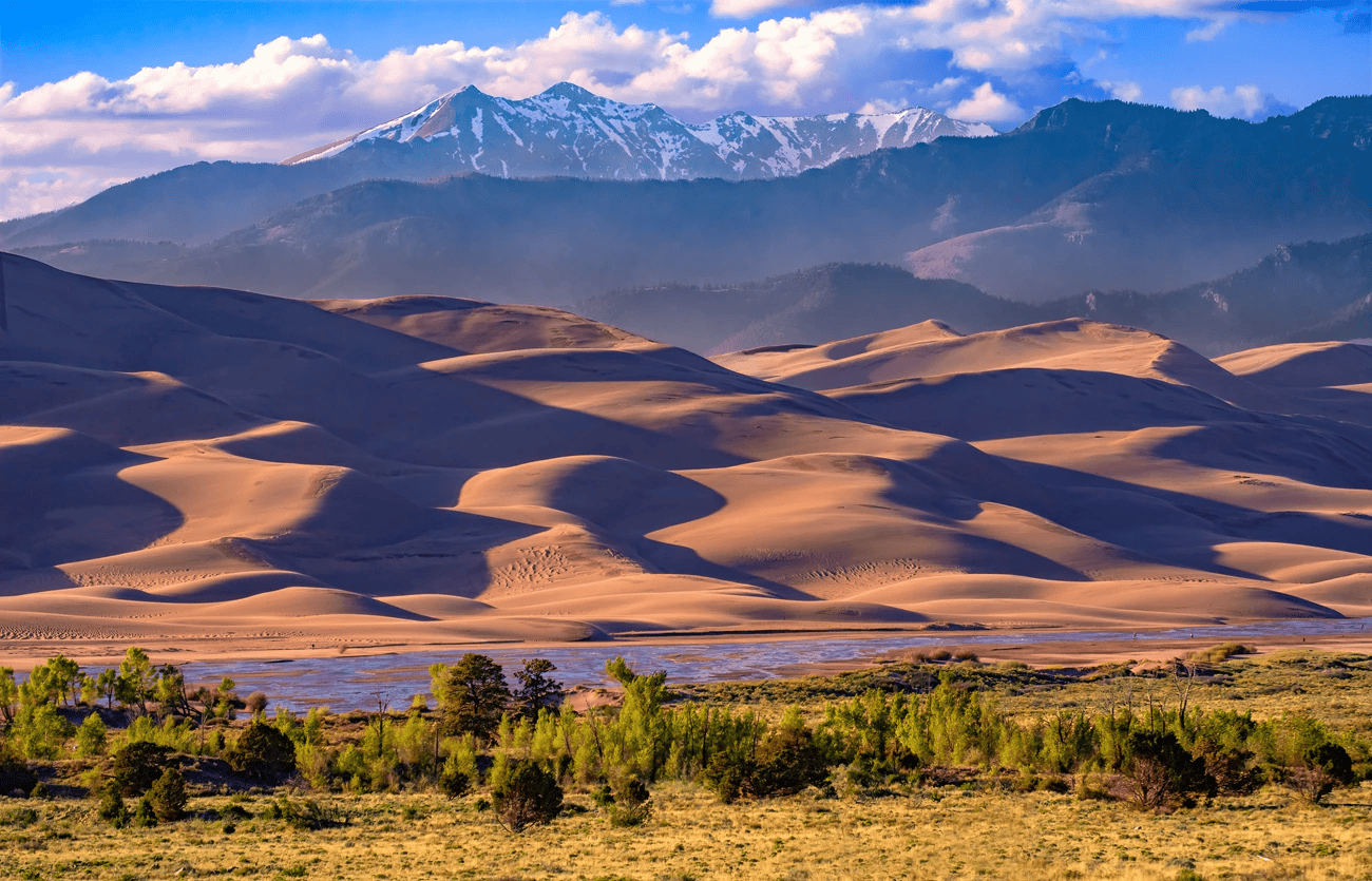 A ground view of the Great San Dunes National Park, complete with the creek at the base, sand dunes behind it, and rows of growing mountains until the final towering row that is dusted in snow.