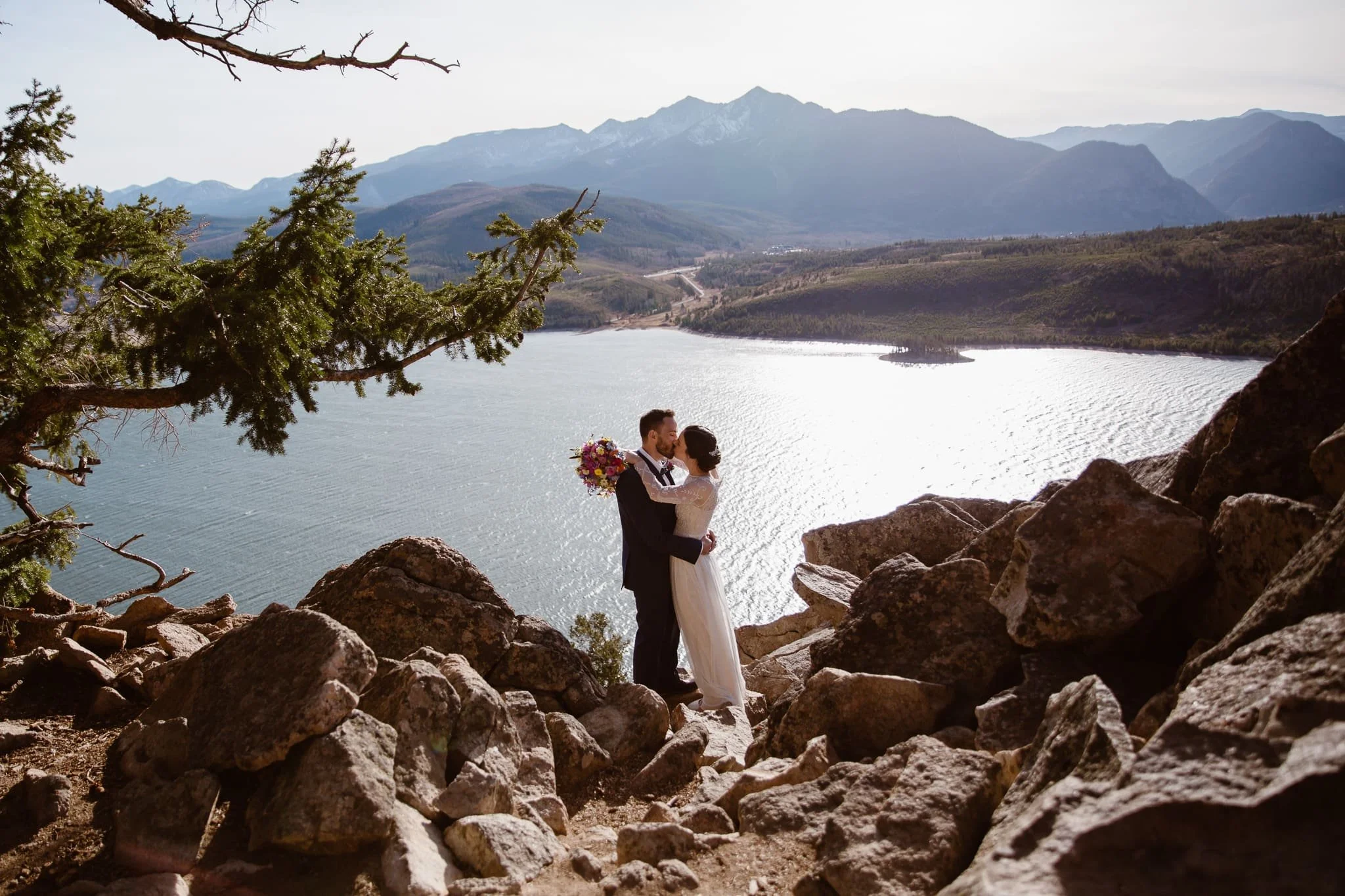 Bride and groom kiss while standing among boulders in front of a large lake with mountains