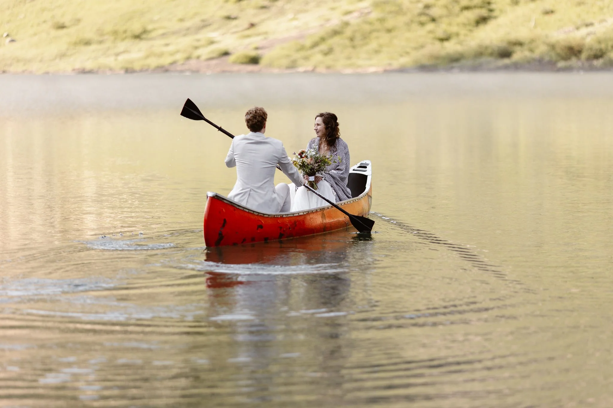 During their sunrise elopement on a lake in Crested Butte, the bride and groom sit in a red canoe as the groom paddles the across the lake, and the bride holds her flowers while wrapped in a cozy blanket.