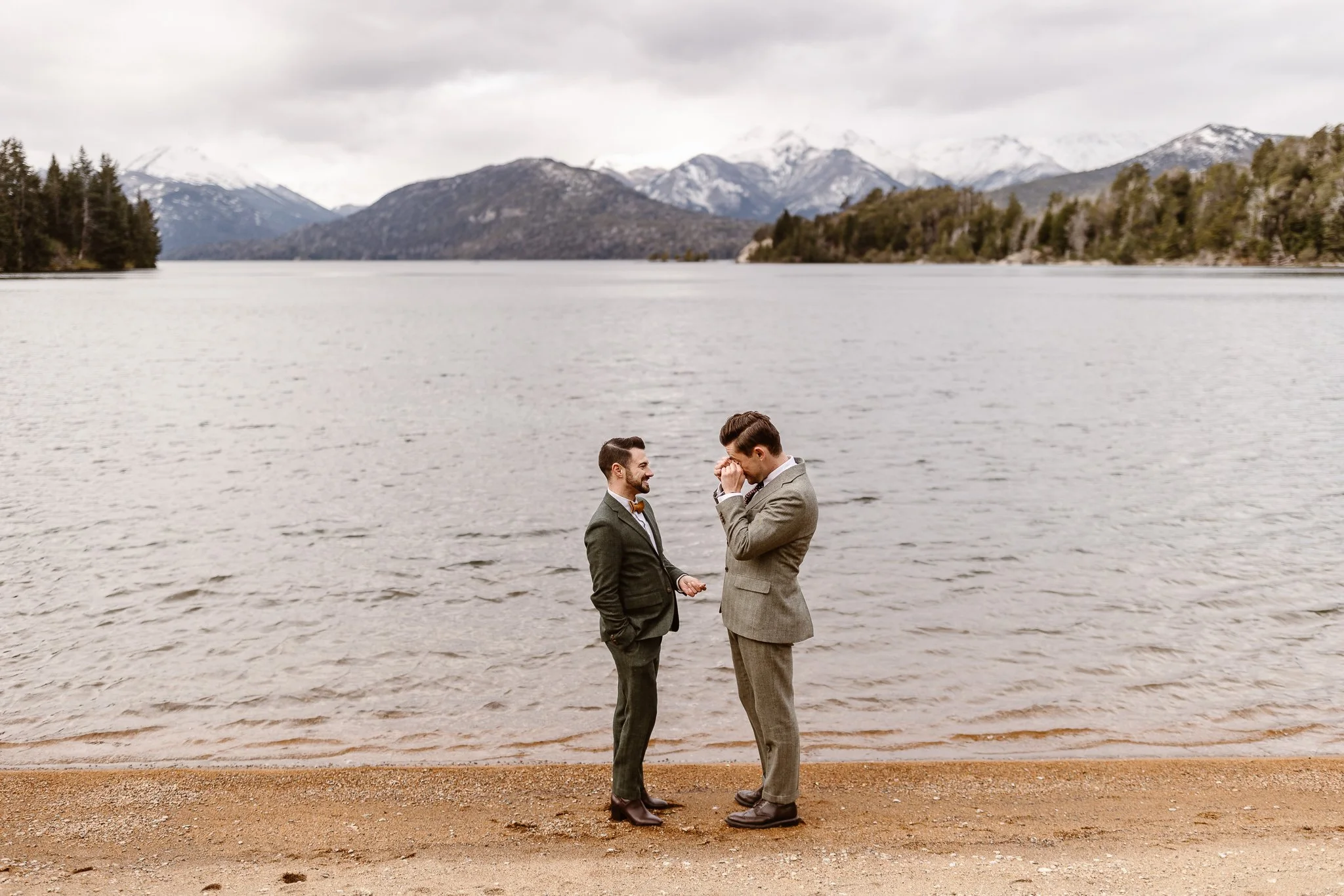 During their afternoon elopement on the shore of vast lake in Bariloche, Patagonia, the grooms share an emotional moment during their private ceremony. The groom on the right is wiping tears from his eyes while the groom on the left smiles sweetly.