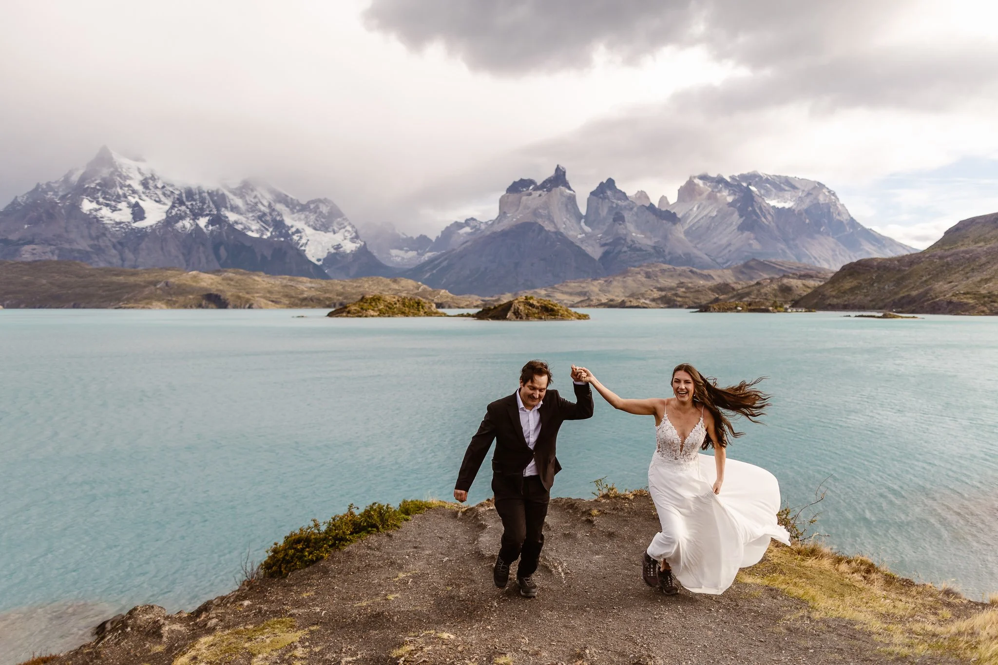 Smiling in celebration of their marriage, the bride and groom hold hands while walking away from the edge of the water in Torres del Paine. Her dress has a lace top and flowy skirt that's blowing in the breeze, while he wears a black suit.