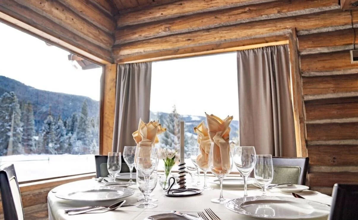 The interior of the Ski Tip Lodge, showing a dining table that is ready for an event, situated in the corner between two huge windows that show the beautiful view of the trees and mountains outside.