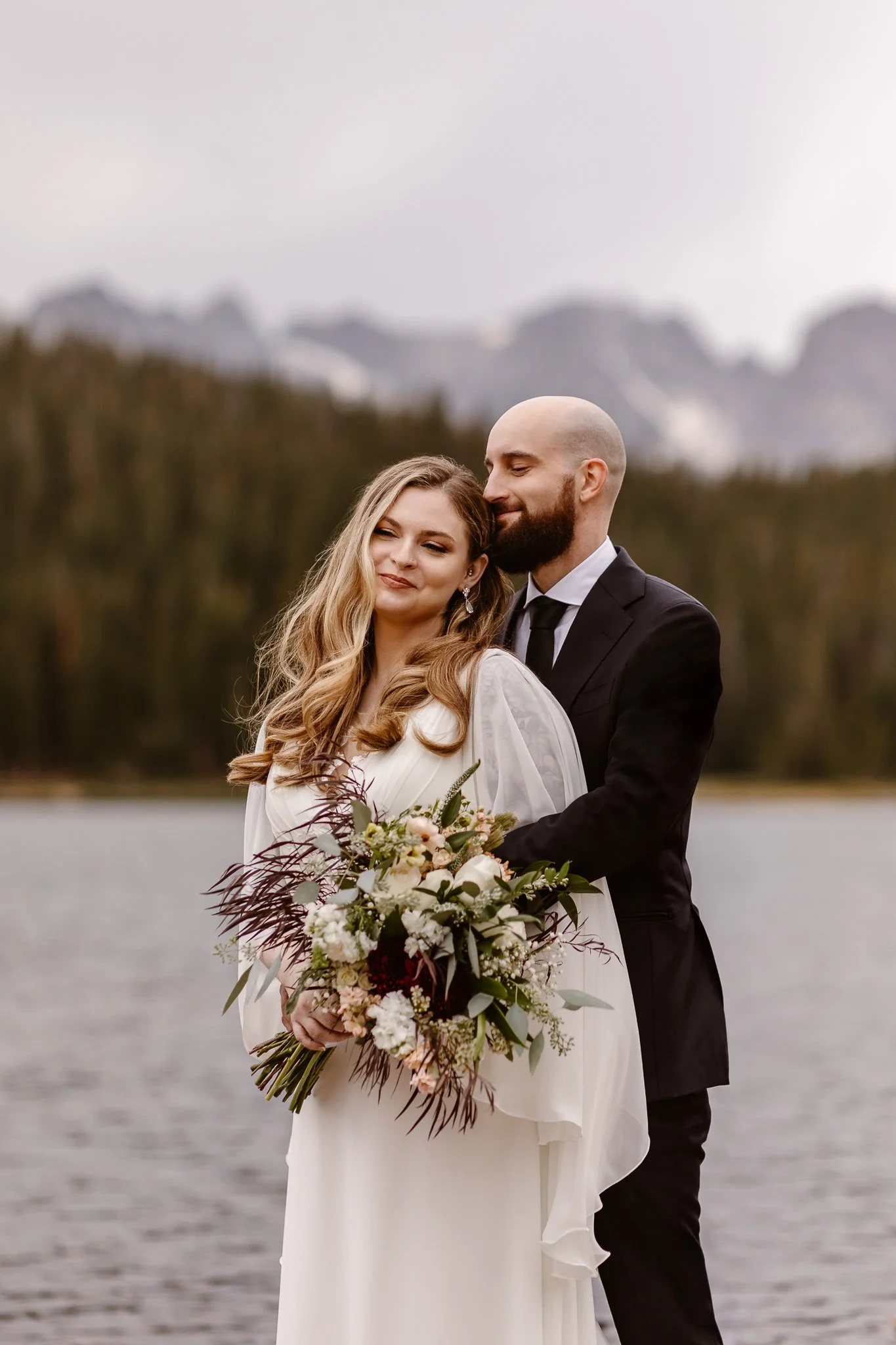 In a gentle moment, the groom hugs his bride from behind with his eyes closed and a sweet smile on his face, as she holds her bouquet and smiles.