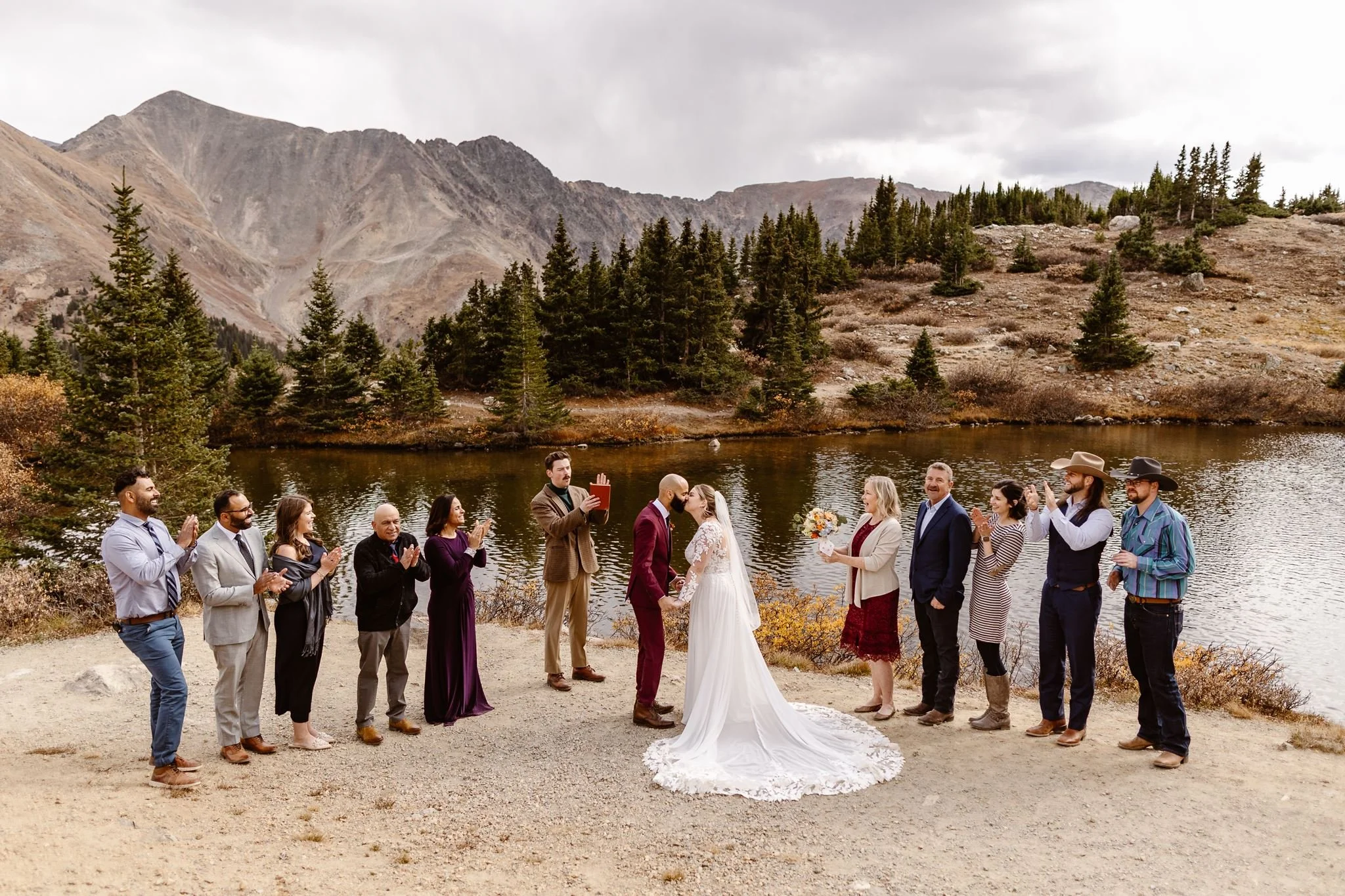 As the bride and groom seal their vows with the first kiss, their families on either side of them clap in celebration during this afternoon micro wedding on the shore of an alpine lake in the heart of Breckenridge, CO.