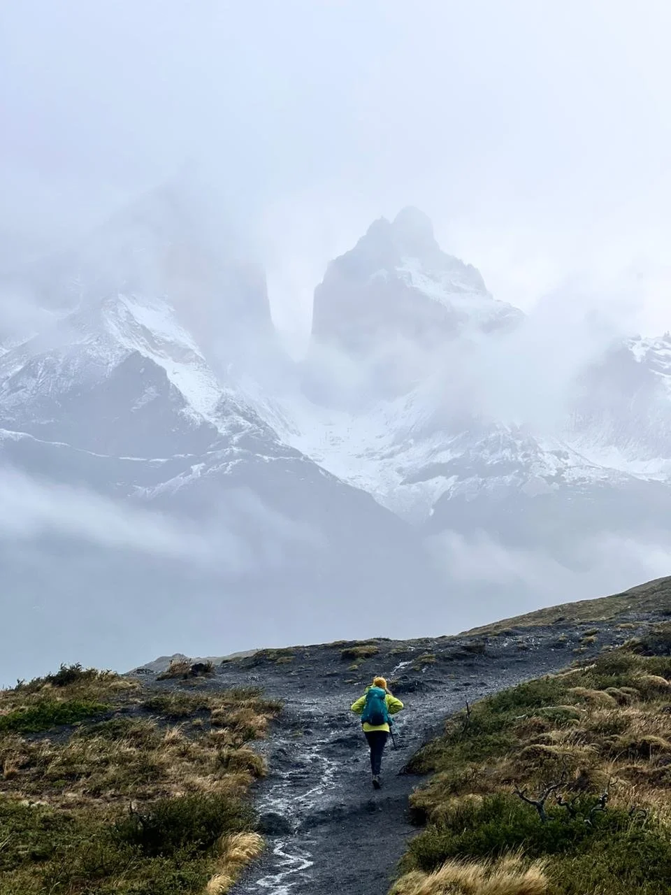 Elopement photographer, Andrea Enger, walking away from the camera with a teal backpack on her back, walking towards the towering peaks of Patagonia on a cloudy and foggy morning.