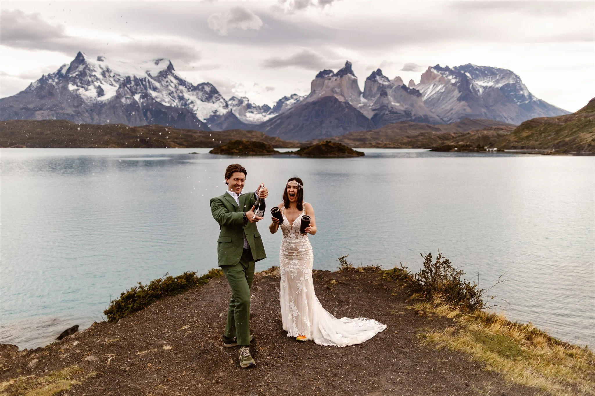 During an afternoon elopement celebration on the water's edge in Torres del Paine, Patagonia, the bride and groom smile joyfully as he pops a bottle of champagne in his green suit and she stands to the right holding custom cups in her beautiful dress