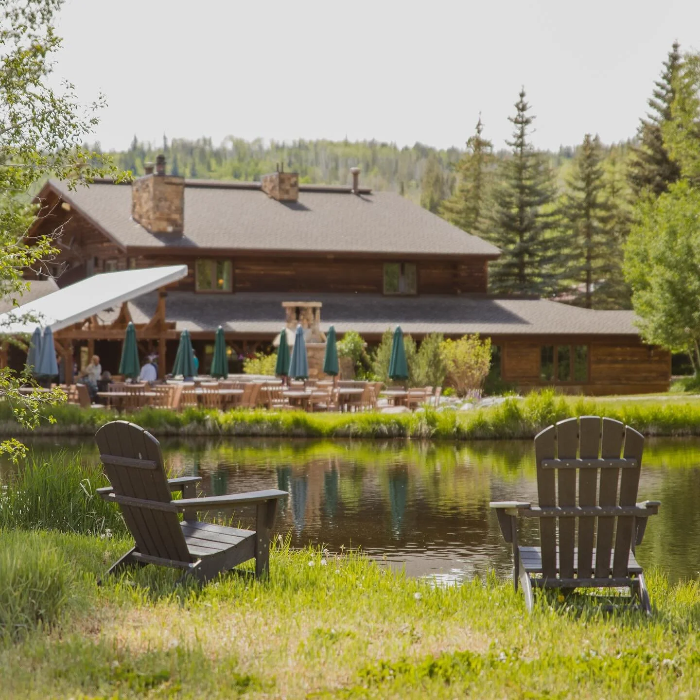 Two wooden chairs face the lake and building at Vista Verde where wedding receptions are held amongst the wooden tables with their blue umbrellas, surrounded by trees and nature.