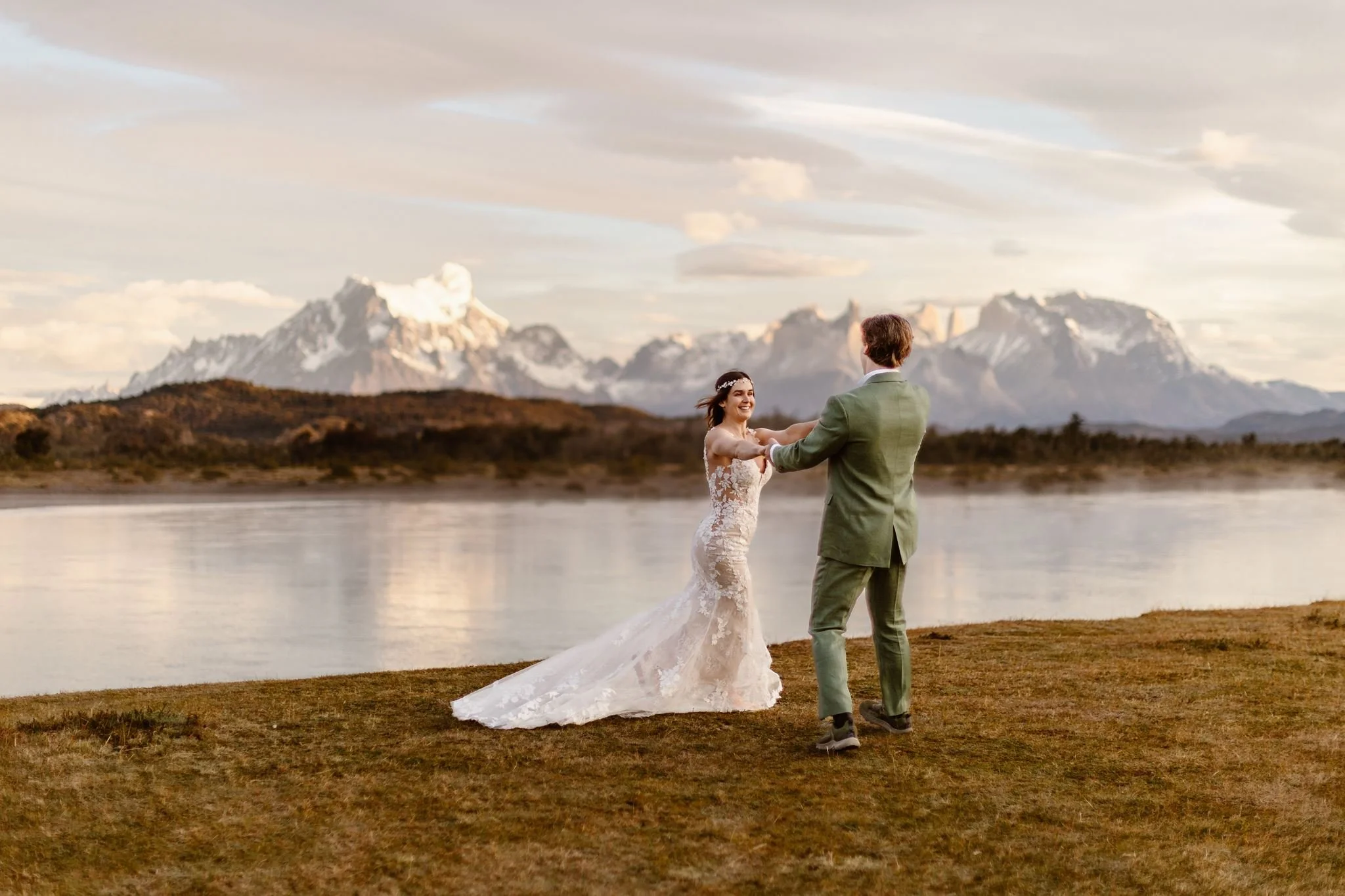 A photo of the bride and groom holding each other's hands while spinning around in a circle, having the time of their lives. They do so on the edge of the water while the sun slowly rises.