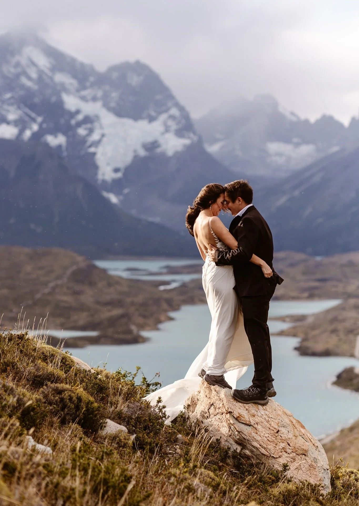 Newlyweds hold each other close while standing on a boulder at the edge of a grand vista with lakes and mountain peaks in Patagonia, Chile.