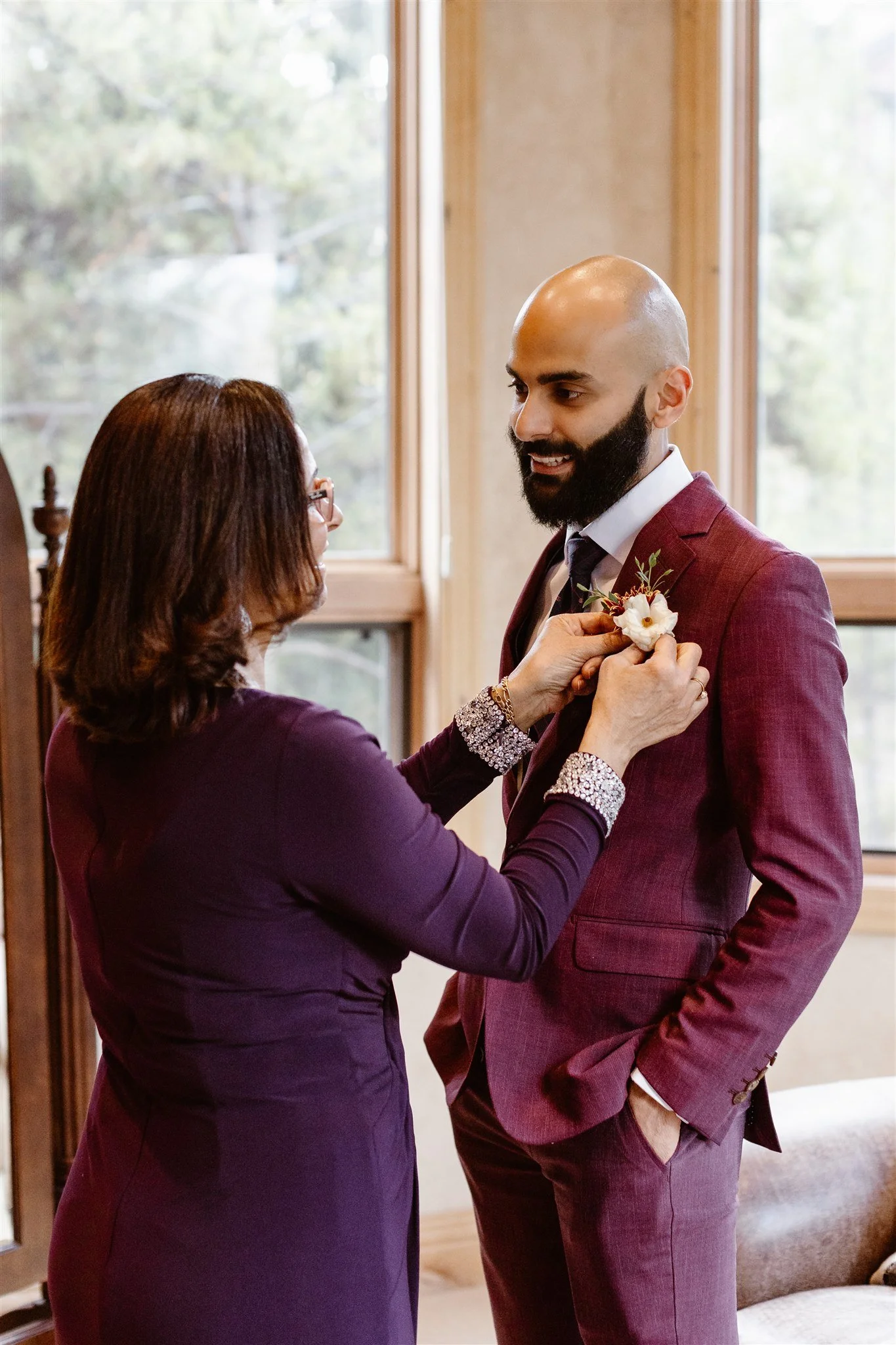 The mother of the groom adding the final touch to her son's wedding suit with a delicate boutonniere made of a single white flower with some green and orange pieces on the left side.
