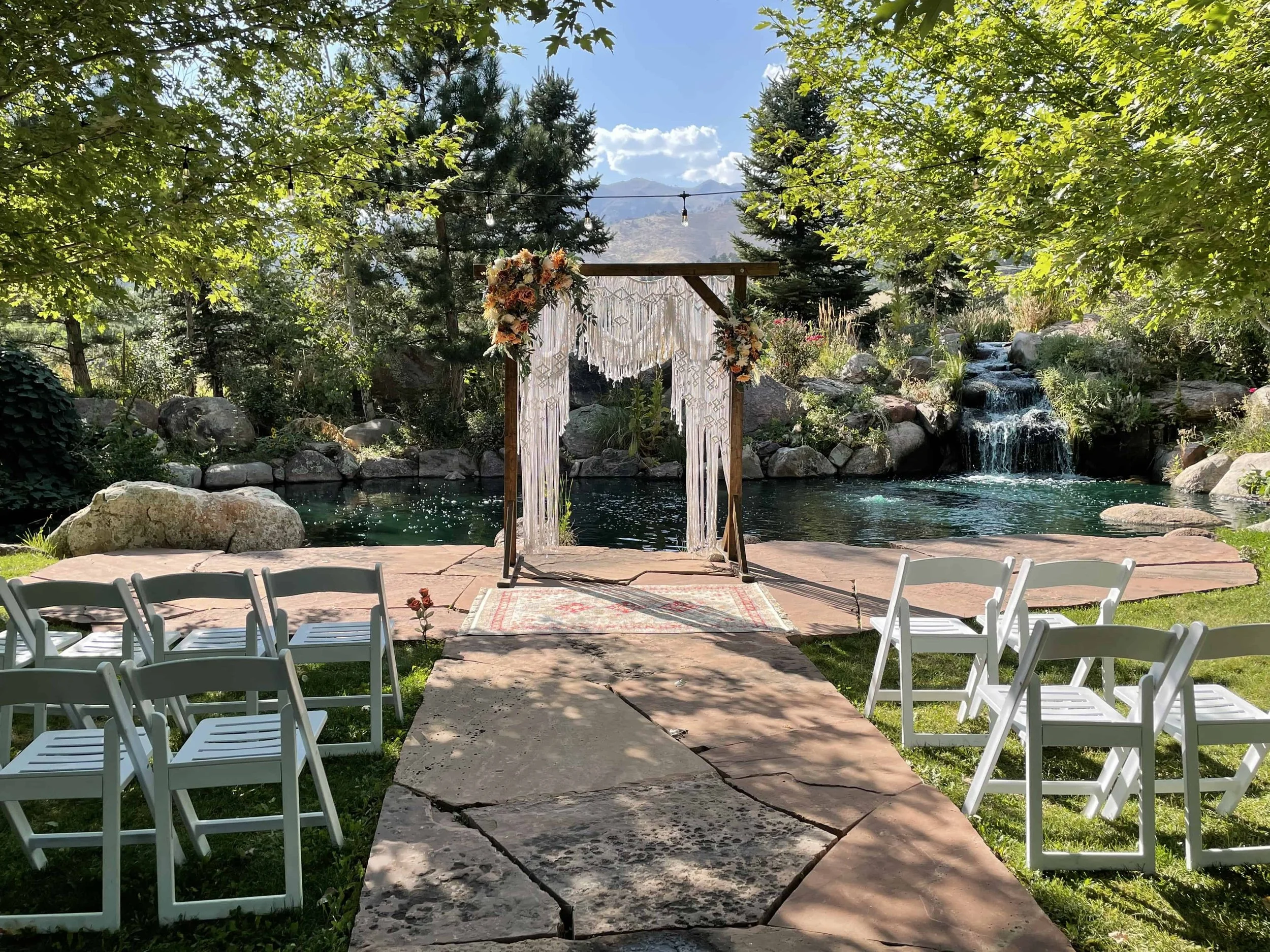 An arch with fringe macrame stands in front of a pond with a dreamy waterfall before a wedding ceremony at Greenbriar Inn. The ceremony site is located on a stone pathway, with white wooden chairs on other side of the path in the grass.