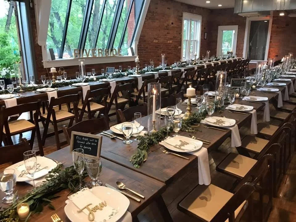 The indoor reception space at The Riverside, set up with 2 long rows of wooden tables and chairs, adorned with candles and simple place settings.