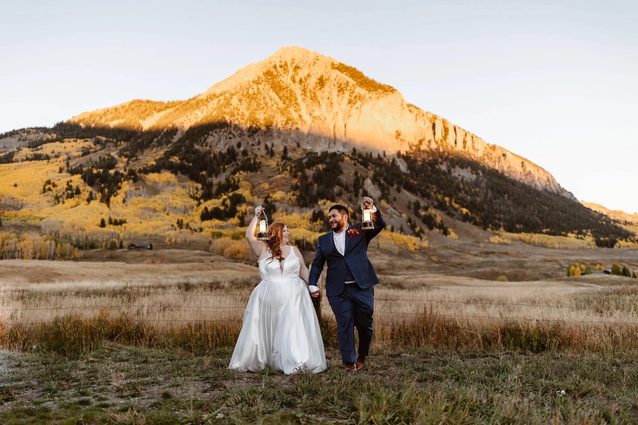 As the golden sunset illuminates the top half of the mountain behind them and the yellow aspen trees fill the ground below, an interracial plus-sized bride and groom hold hands and lanterns in the air, while smiling sweetly at each other.