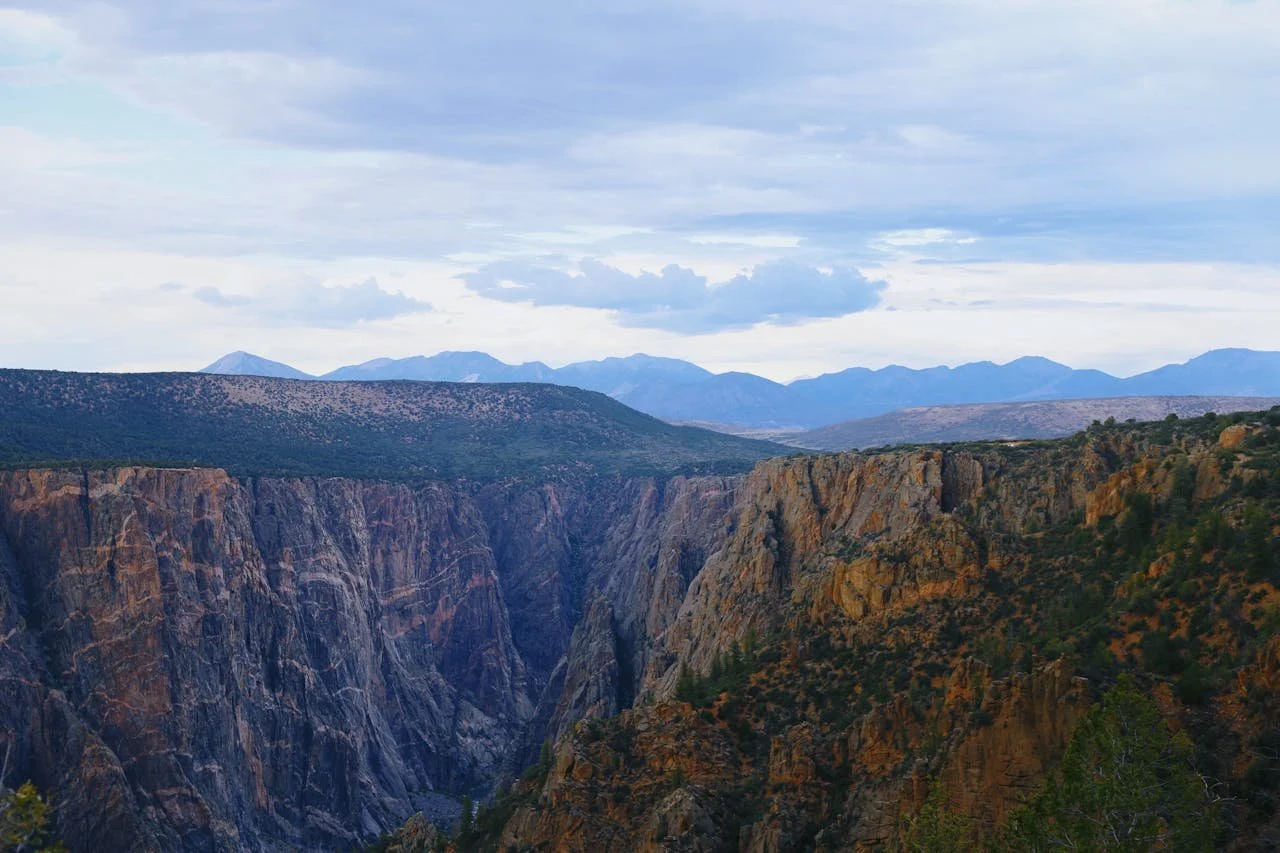 A view from the top of the Black Canyon of the Gunnison National Park. You can see part of the marbled gorge below, the flat land the rims the canyon, and the mountains that fill the background on a cloudy afternoon.
