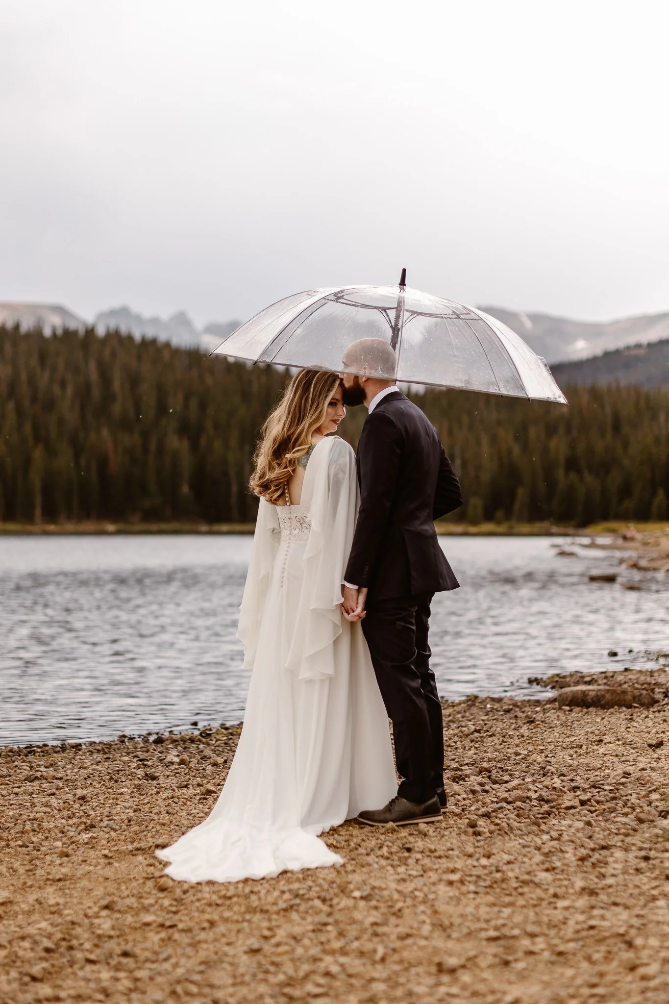 On the shore Brainard Lake, the eloping couple hold hands under a clear umbrella that is being held by the groom. As he kisses the bride's forehead, she looks towards the camera with a gentle smile.
