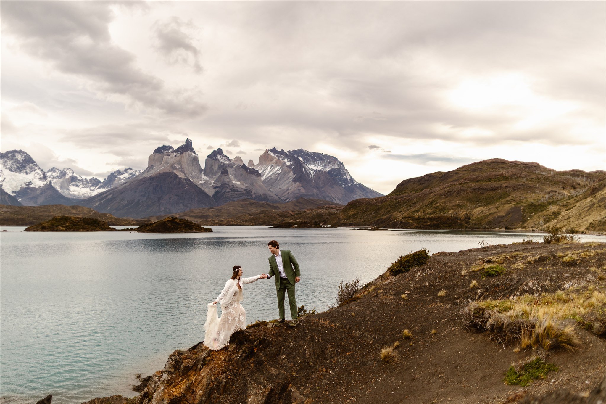 As the newlyweds travel throughout the area, they were climbing a dirt mound and the groom holds her hand as she holds his with her left hand and the train of her dress with the right hand. Behind them the scenery is beautiful and wildly contrasting