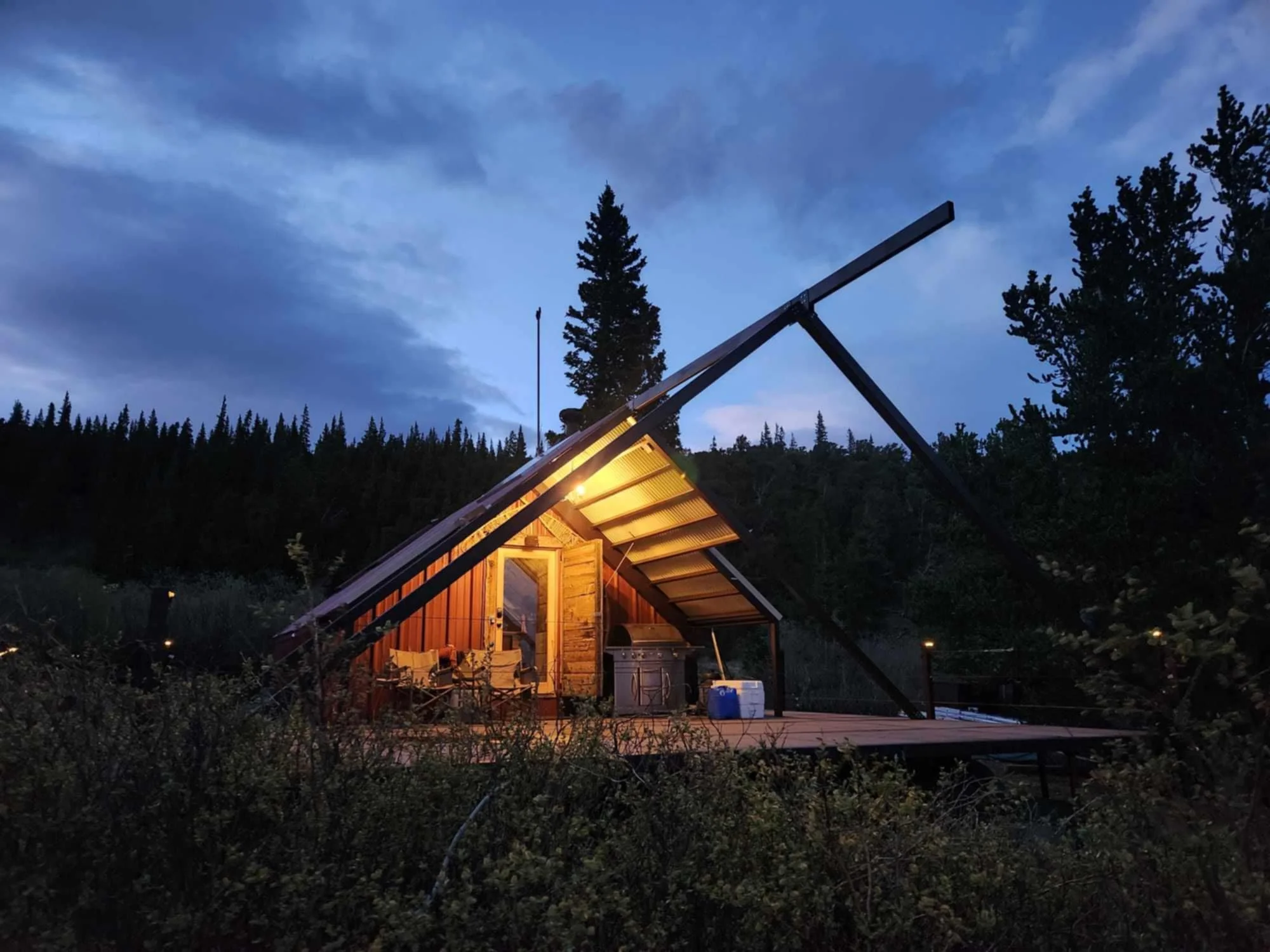 The quaint A-frame hut sits secluded in the night as the singular light glows from the roof, illuminating the wooden entryway and deck below.