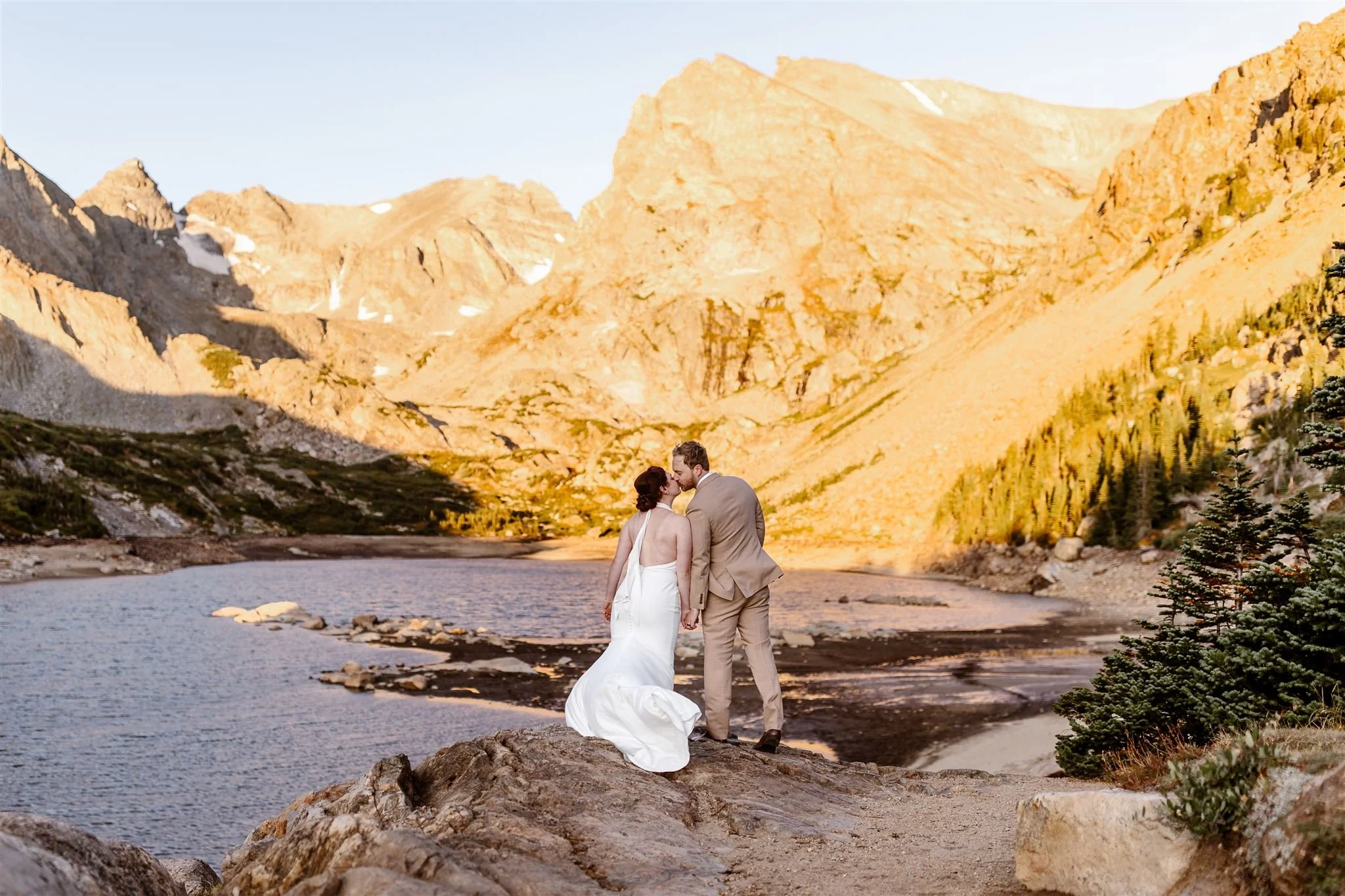 On a September morning at Lake Isabelle, the bride and groom stand side-by-side, hand-in-hand, as they share a beautiful kiss during a stunning orange sunrise on the mountains surrounding the lake. The water ripples before them as the wind picks up.