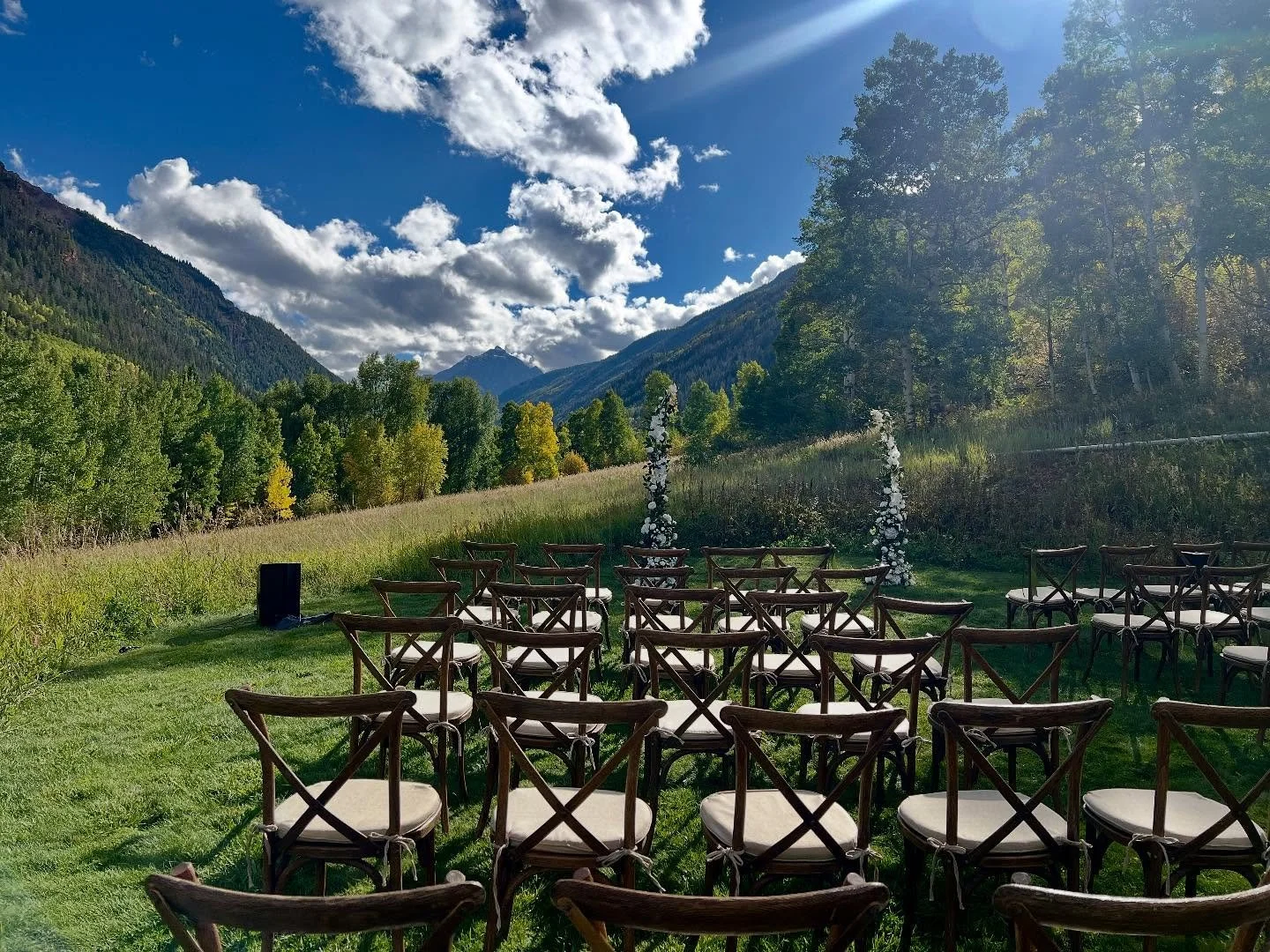 The view from the seating in the grass at T-Lazy 7 ranch where a wedding ceremony is set up in a clearing that's surrounded by trees, mountains, and blue skies.