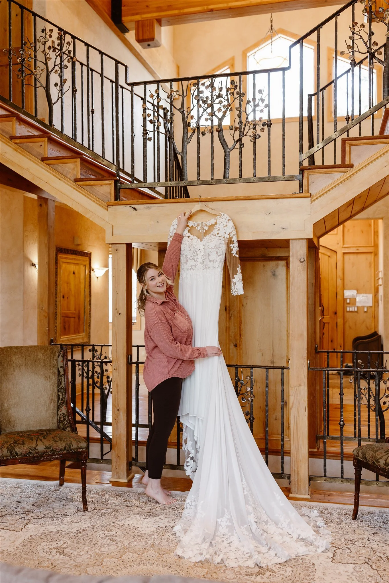 The bride reaches up to grab her wedding dress as it hangs from the stairwell in their cabin-esque rental home. The dress is a long sleeve a-line gone with sheer lace sleeves as the long lace-trimmed train reaches the floor.