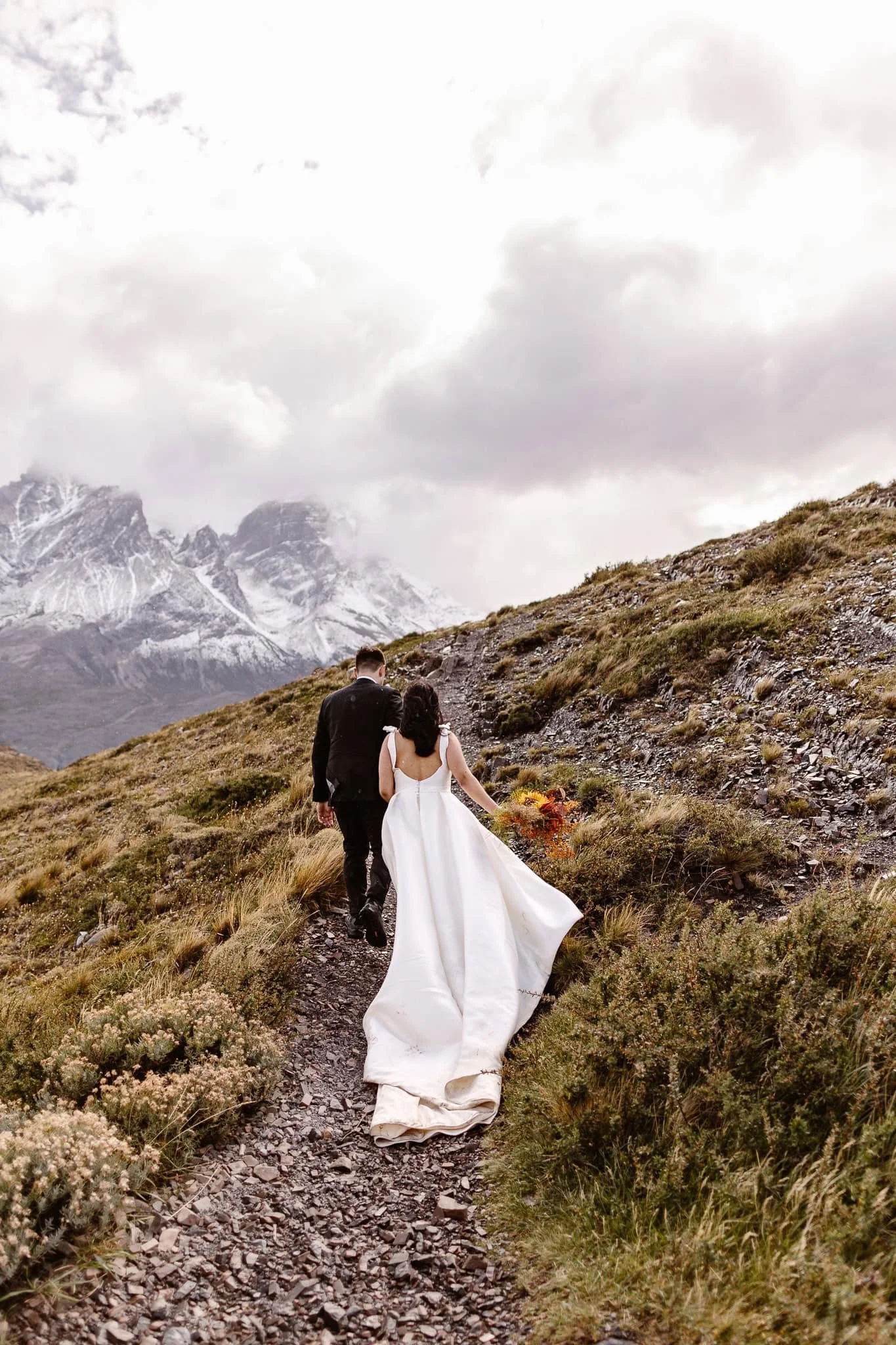 A bride and groom hike up a rocky route toward snow dusted peaks in Patagonia Chile
