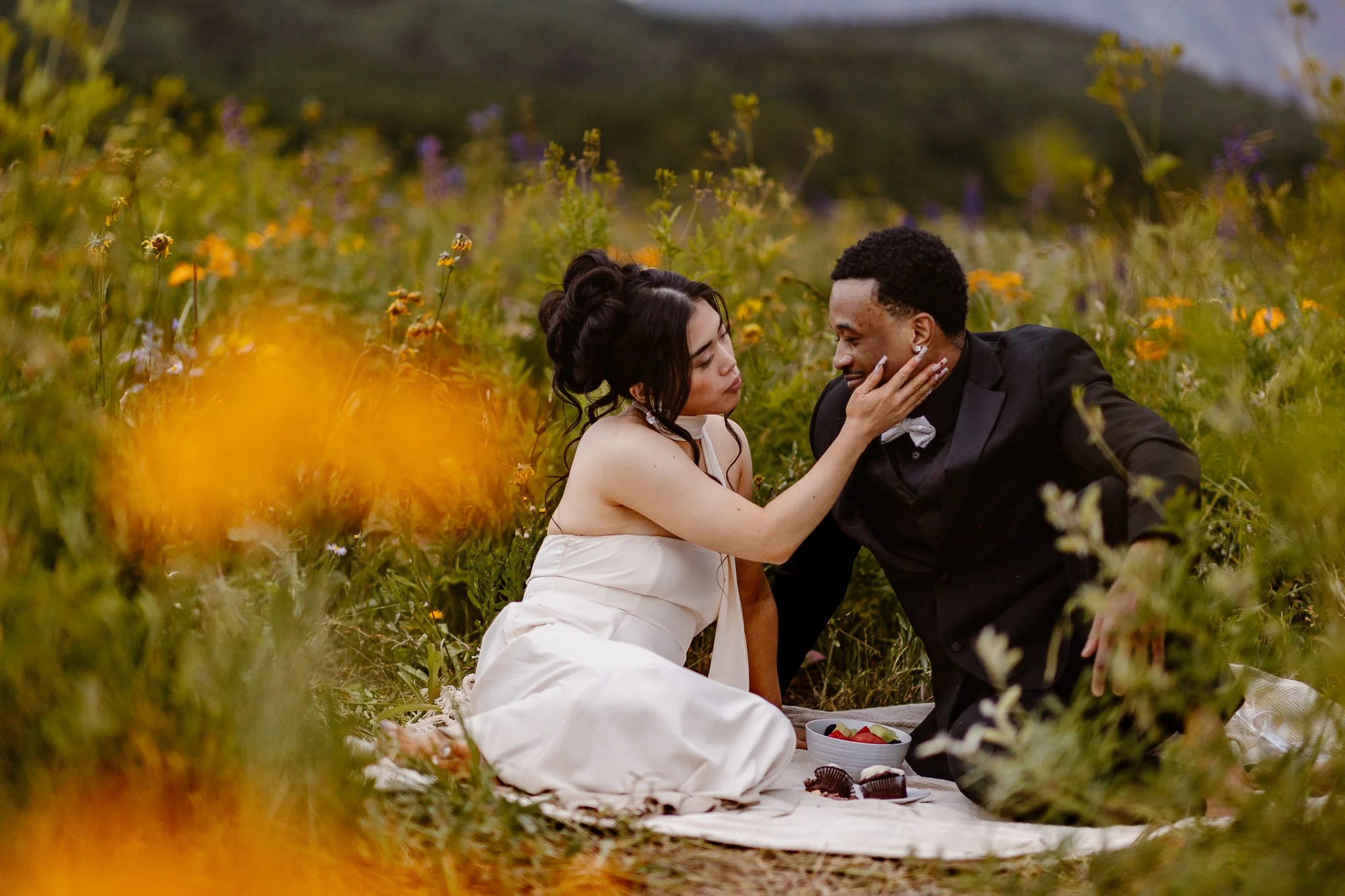 During their elopement picnic, an interracial couple share a tender moment as the yellow flowers hold witness to their love. The two are looking deeply into each other's eyes as the bride softly rests her right hand on their groom's cheek.