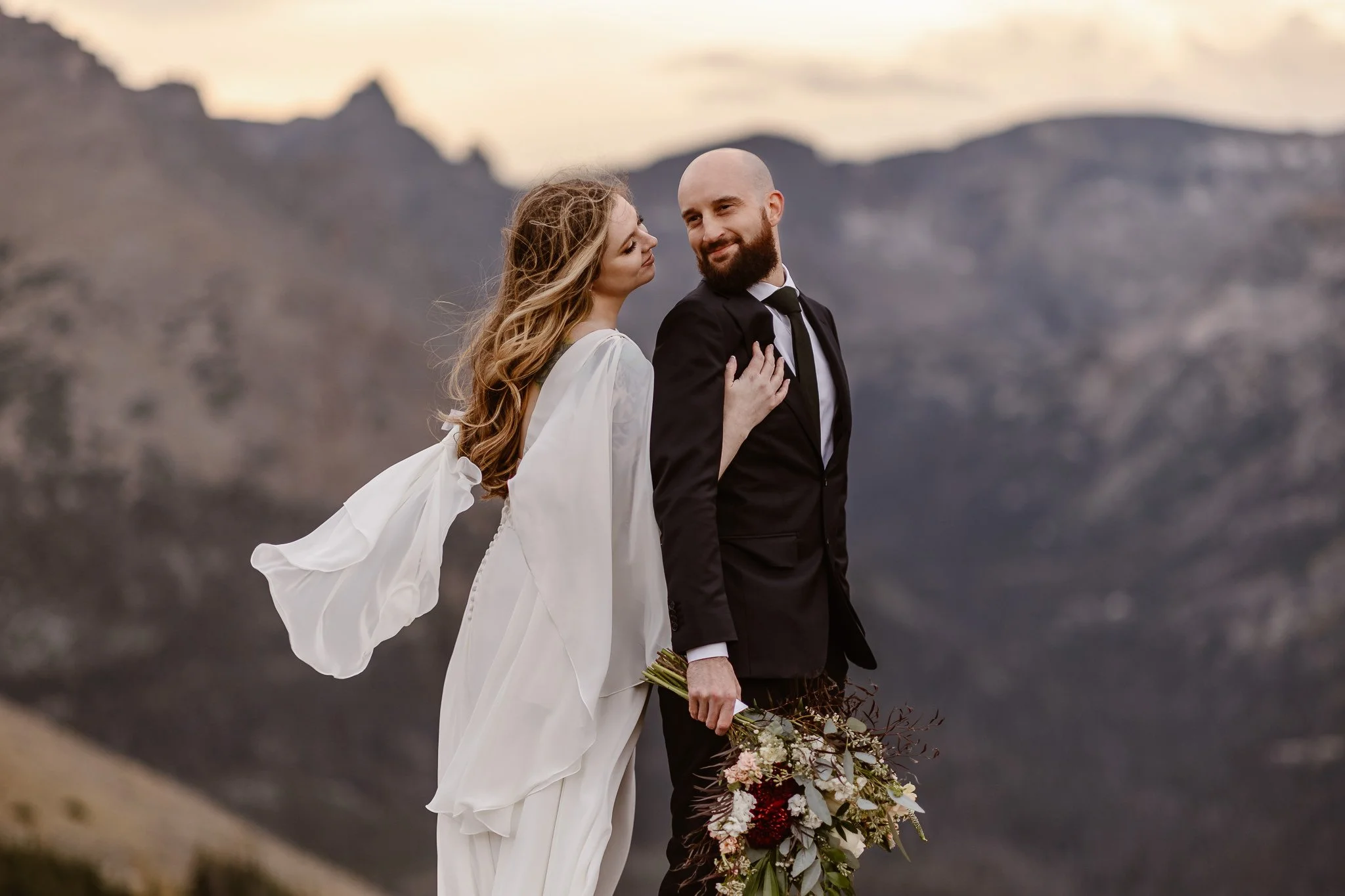 With the grey mountains filling the background, a bride embraces her groom with her right hand and they smile happily in the foreground, the wind blowing the bride's hair and dress.