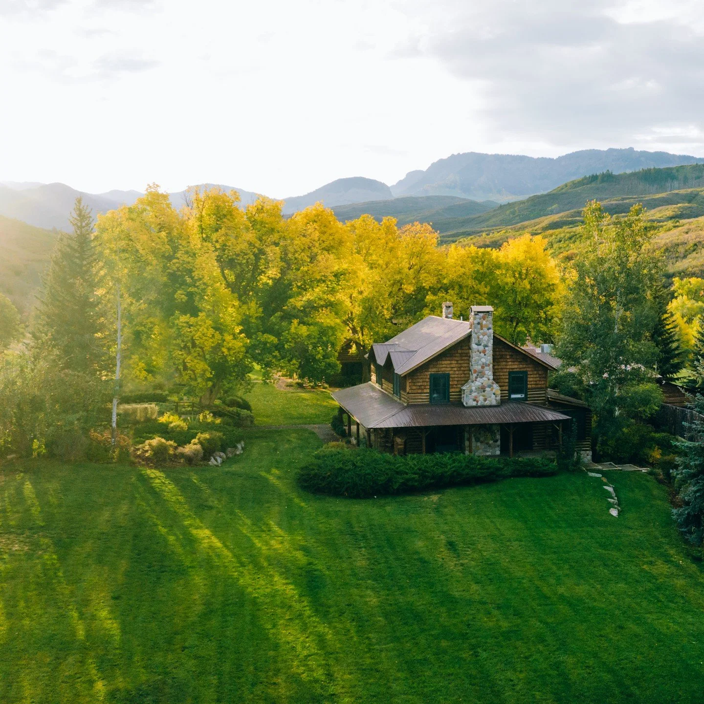 As the sun sets on Smith Fork Ranch, the green and yellow trees illuminate behind the wood and stone building on the lush green lawn below.