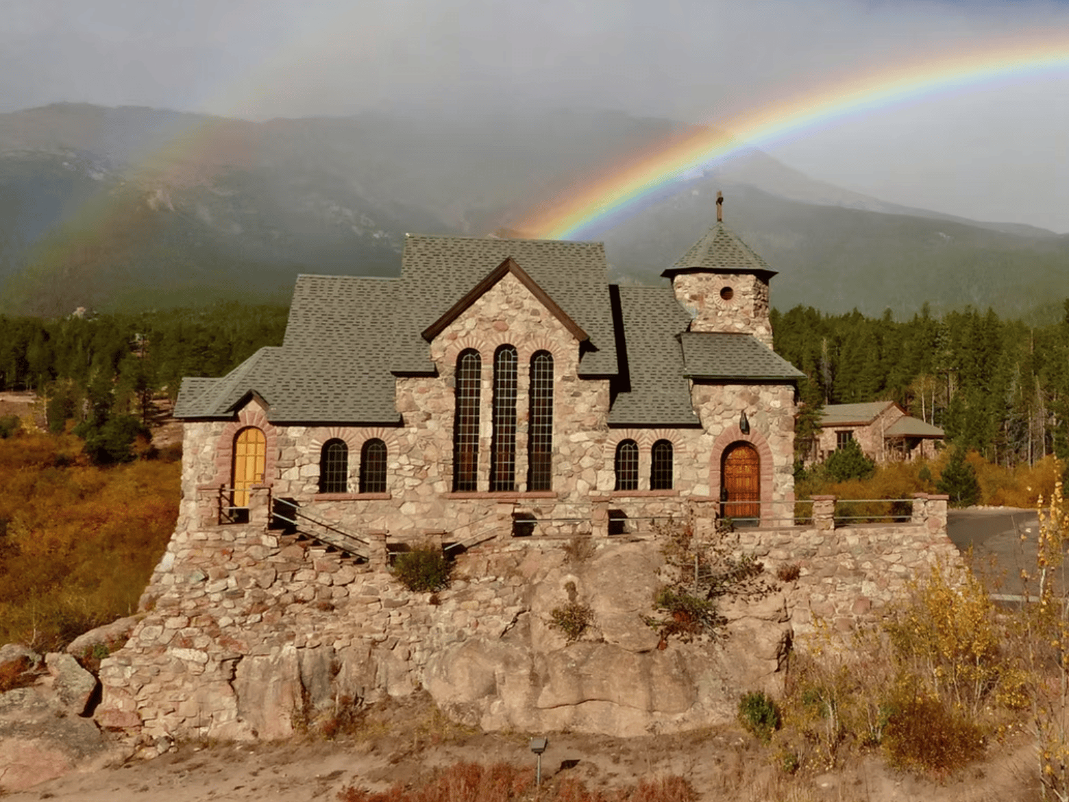 Two rainbows cover the mountains in the background of this exterior photo of Chapel on the Rock at St. Catherine's Chapel. It is a small stone building with tall windows, and wooden doors, that is built atop a large stone formation.