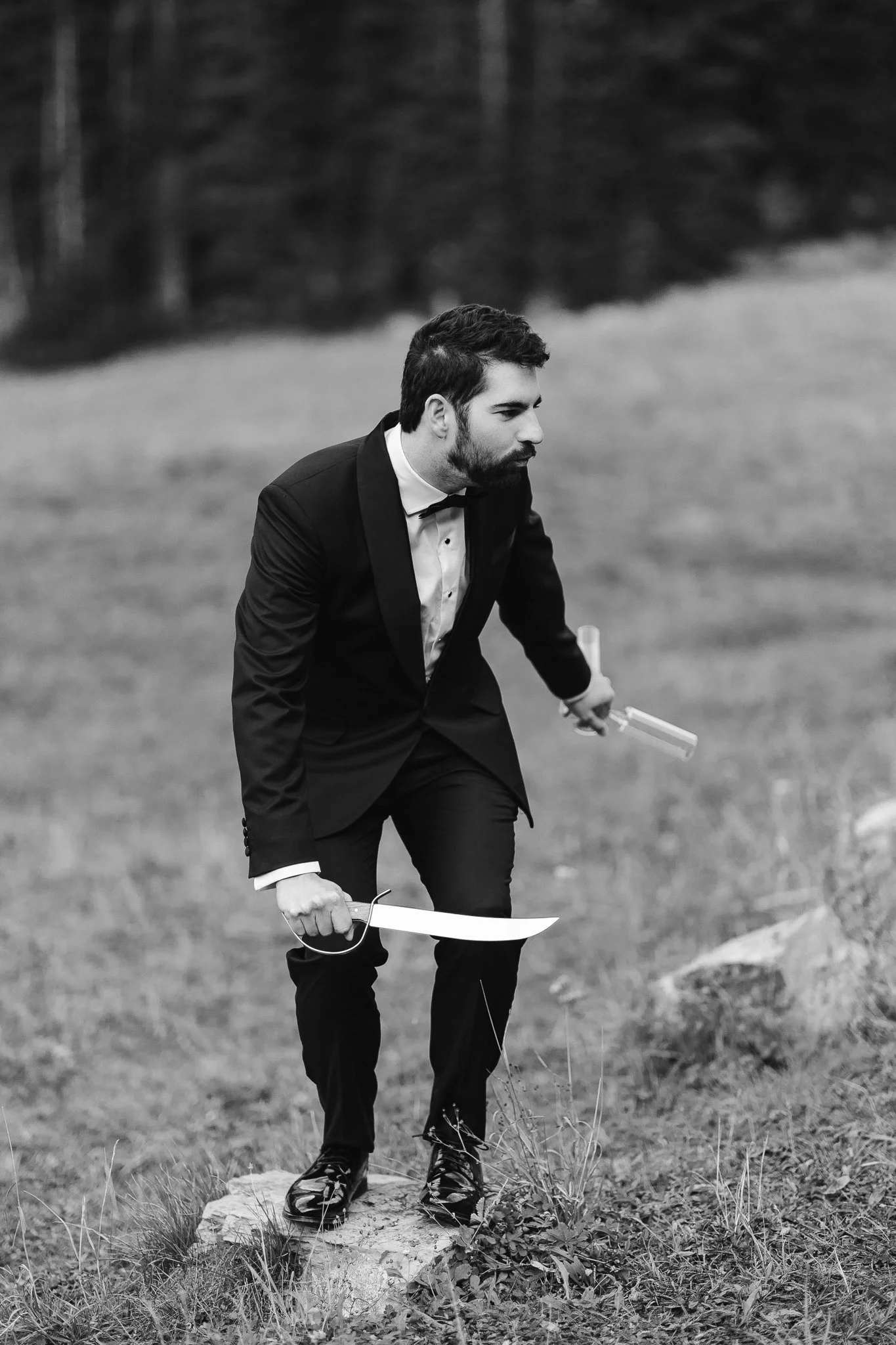 The groom stands on a rock with a saber sword in one hand and two champagne glasses in the other. He is slightly kneeling down with a look that says he's about to go hunting.