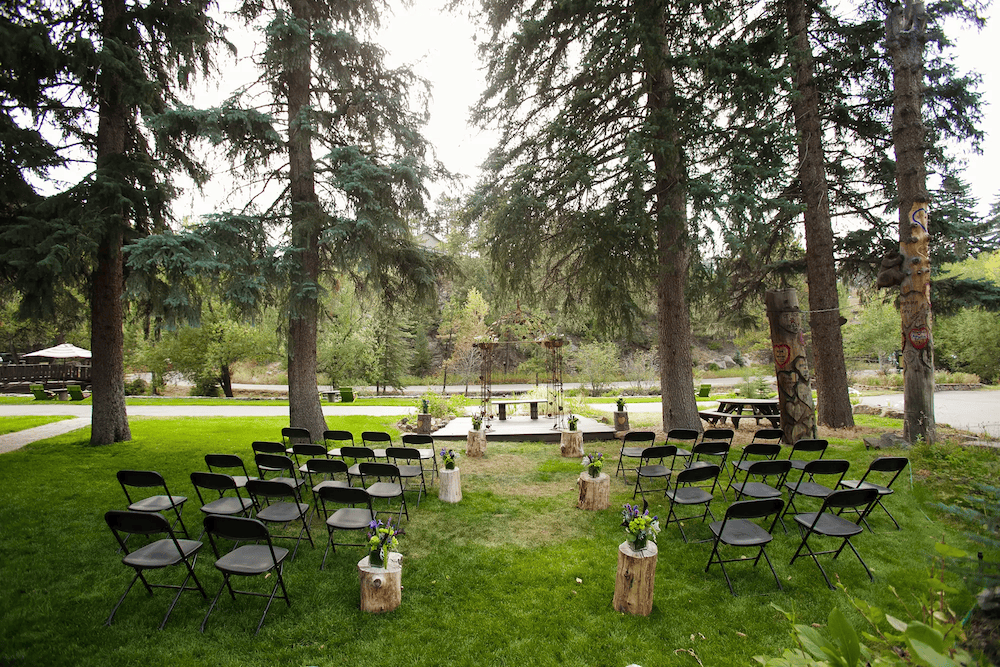 A ceremony set up on the lawn of Highland Haven, with black folding chairs on either side of the aisle that is decorated with short logs and small floral arrangements. There's a metal arch on the platform for the ceremony and behind it all is a creek