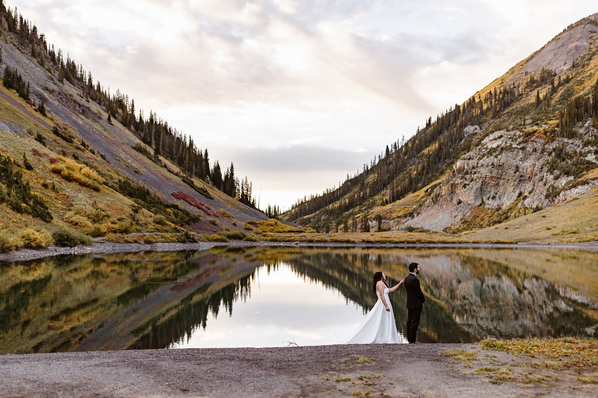 During their sunrise first look at a mirror-like lake with mountains rising to each side from the middle, the groom awaits his bride to the right of the lake as she approaches and taps her grooms shoulder.