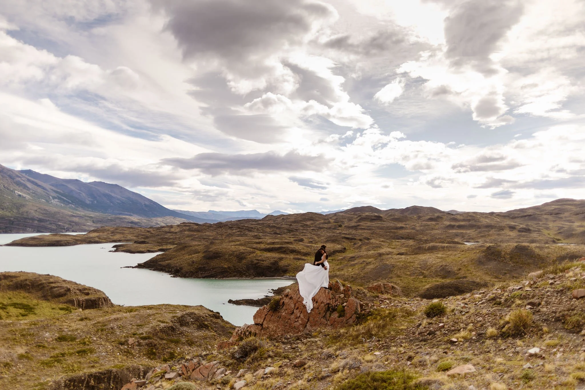 A bride and groom embrace atop a rocky outcrop overlooking a lake and mountains during their Patagonia elopement