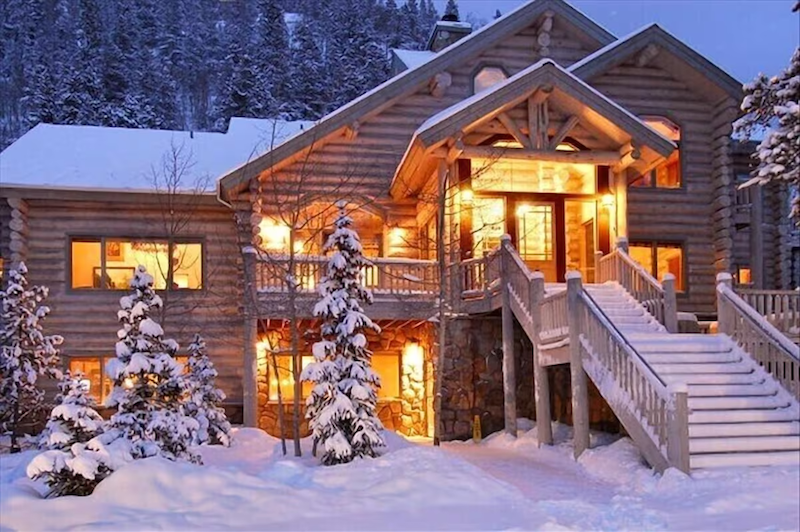 Covered in snow, the Little Mountain Lodge glows in yellow/orange lights that shine from the interior. This cozy lodge has stairs leading up to the log-cabin lodge for a rustic vibe.