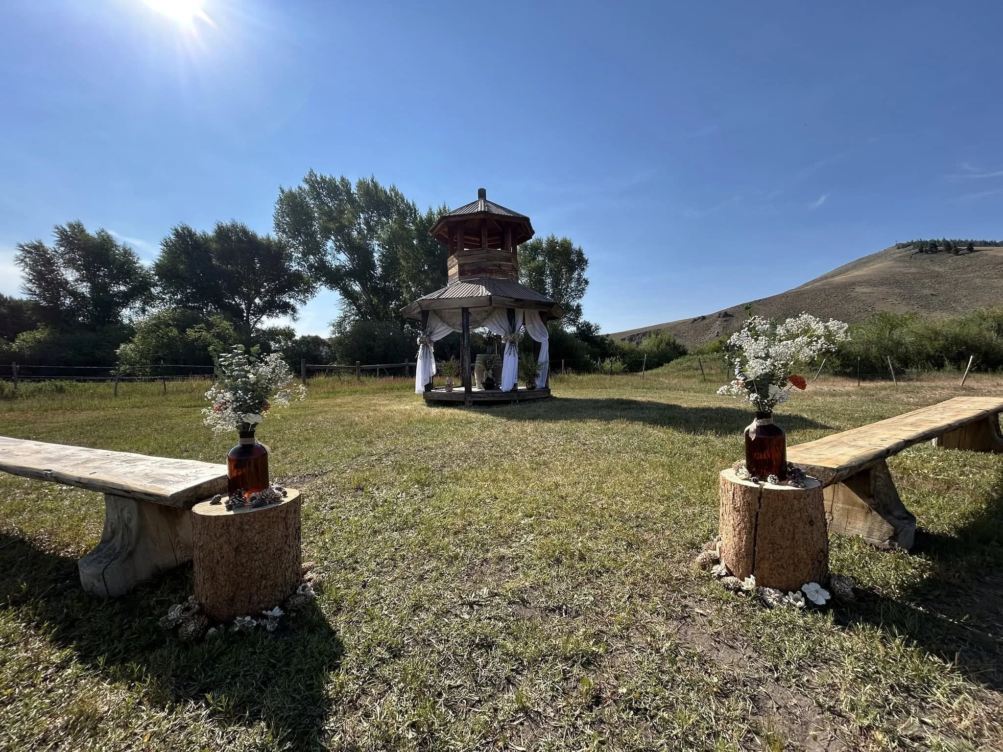 On a sunny afternoon, a wedding ceremony is set up on the open grass area of I Bar Ranch, with wooden benches set up on either side of the aisle, flower vases on the logs along the aisle, and a draped gazebo for the ceremony ahead.