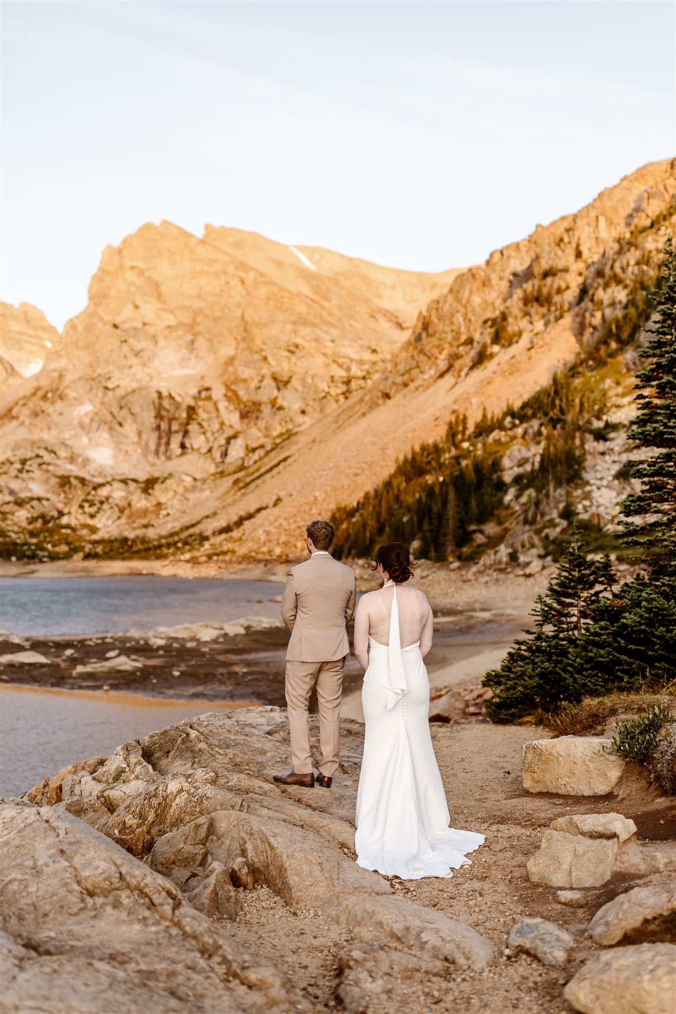 During their sunrise first look on Lake Isabelle, the groom, in a tan suit, stands away from the camera, facing the orange-yellow mountains and lake, while his bride walks up behind him to reveal her gorgeous open-back sheath dress.